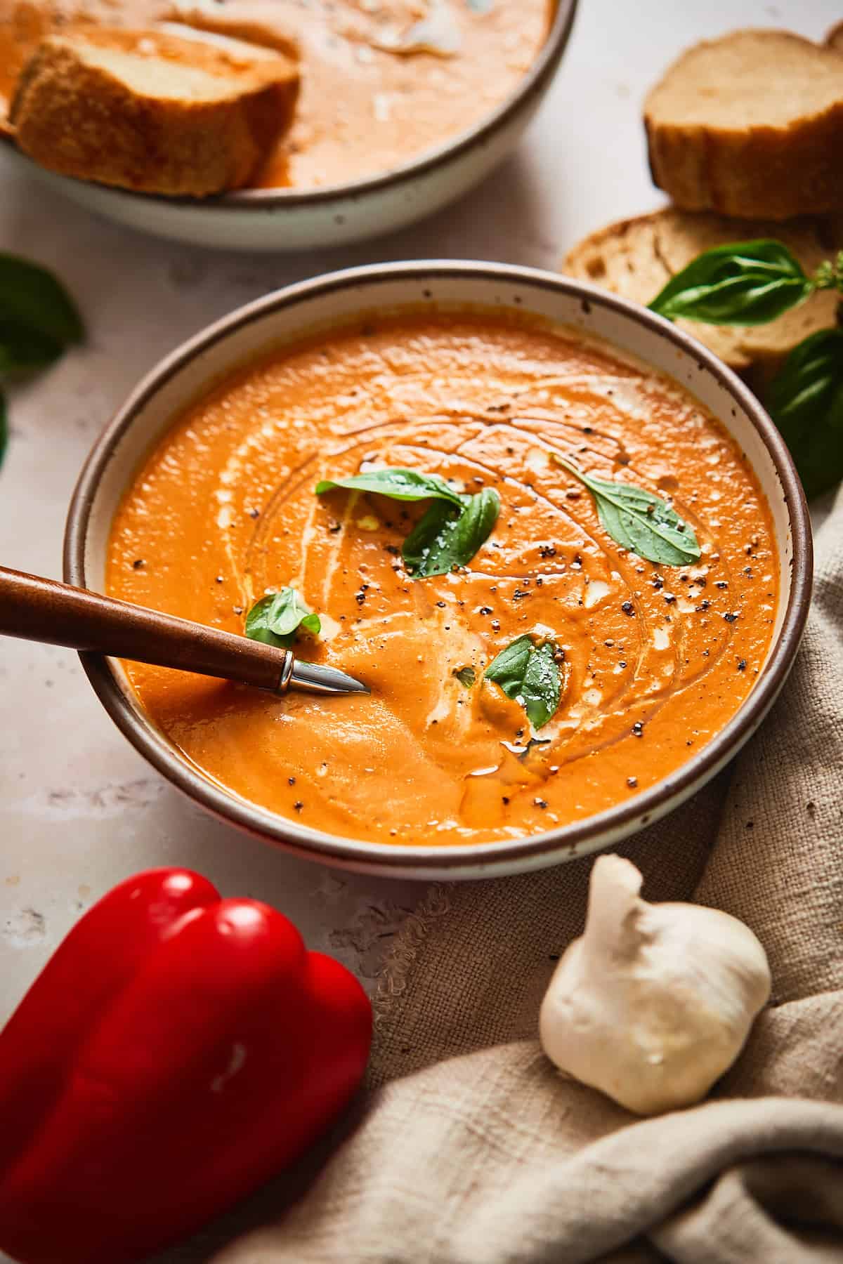 Lovely bowl of roasted tomato and red pepper soup topped with basil, and a spoon in the bowl, surrounded by ingredients used to make the soup. 