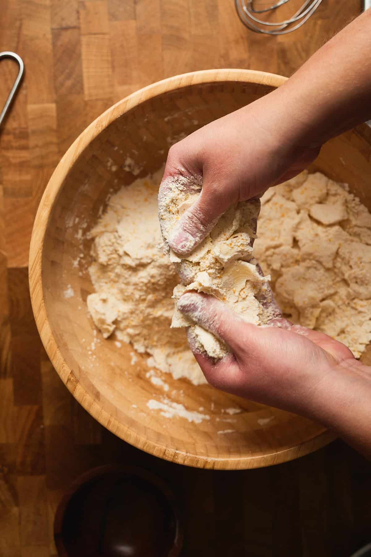 Fresh homemade dough being kneaded in a wooden bowl, with hands pressing the butter into pieces to flatten them.
