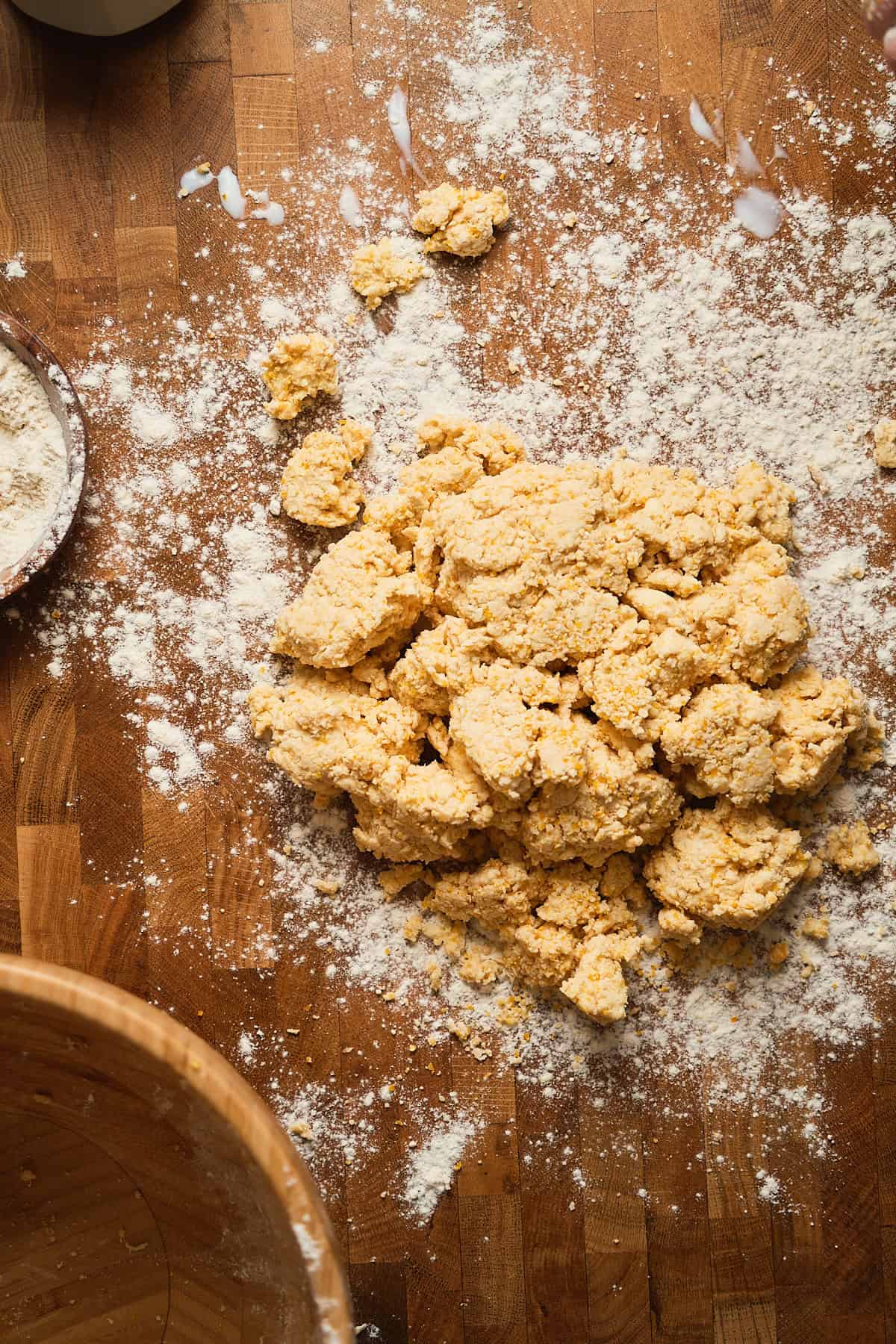 Crumbly cornmeal biscuit dough on wooden surface with scattered flour and dough pieces.