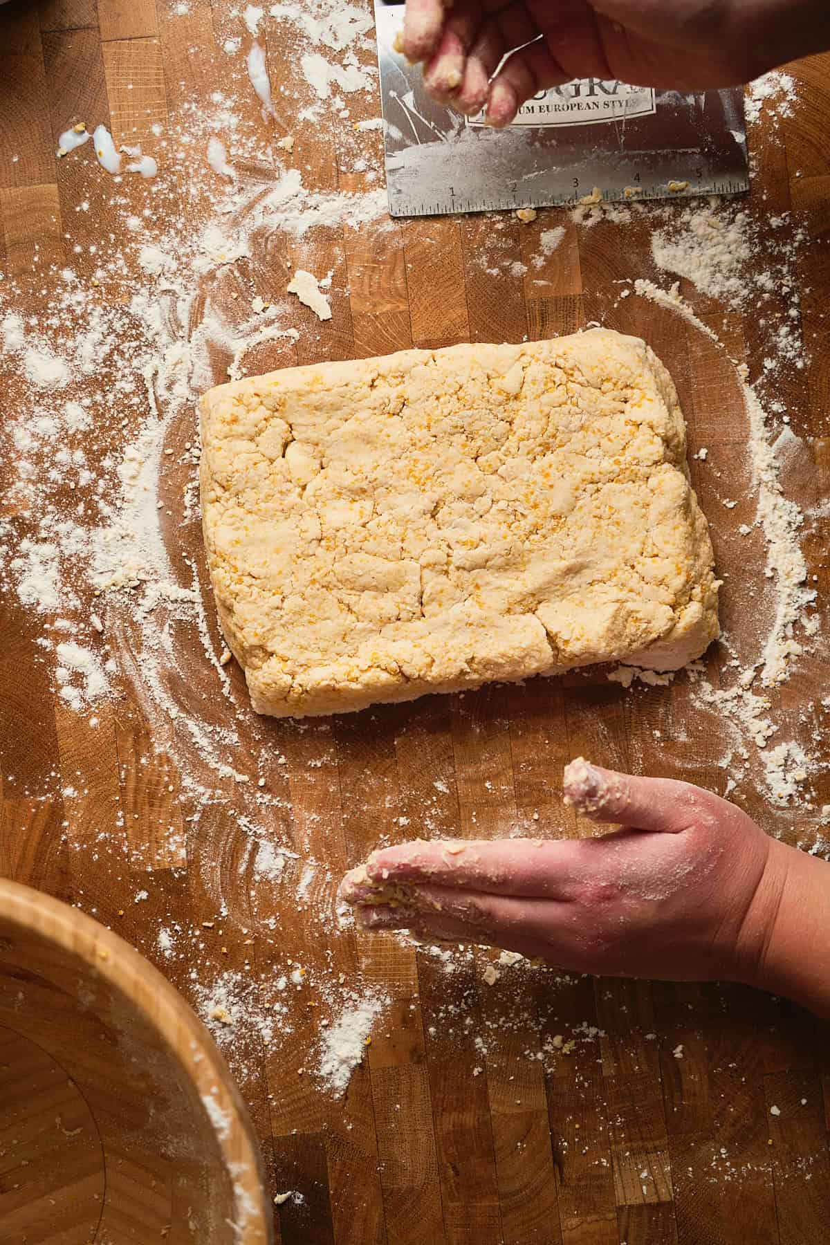 Flaky biscuit dough on wooden surface with hands preparing pastry, surrounded by baking tools.