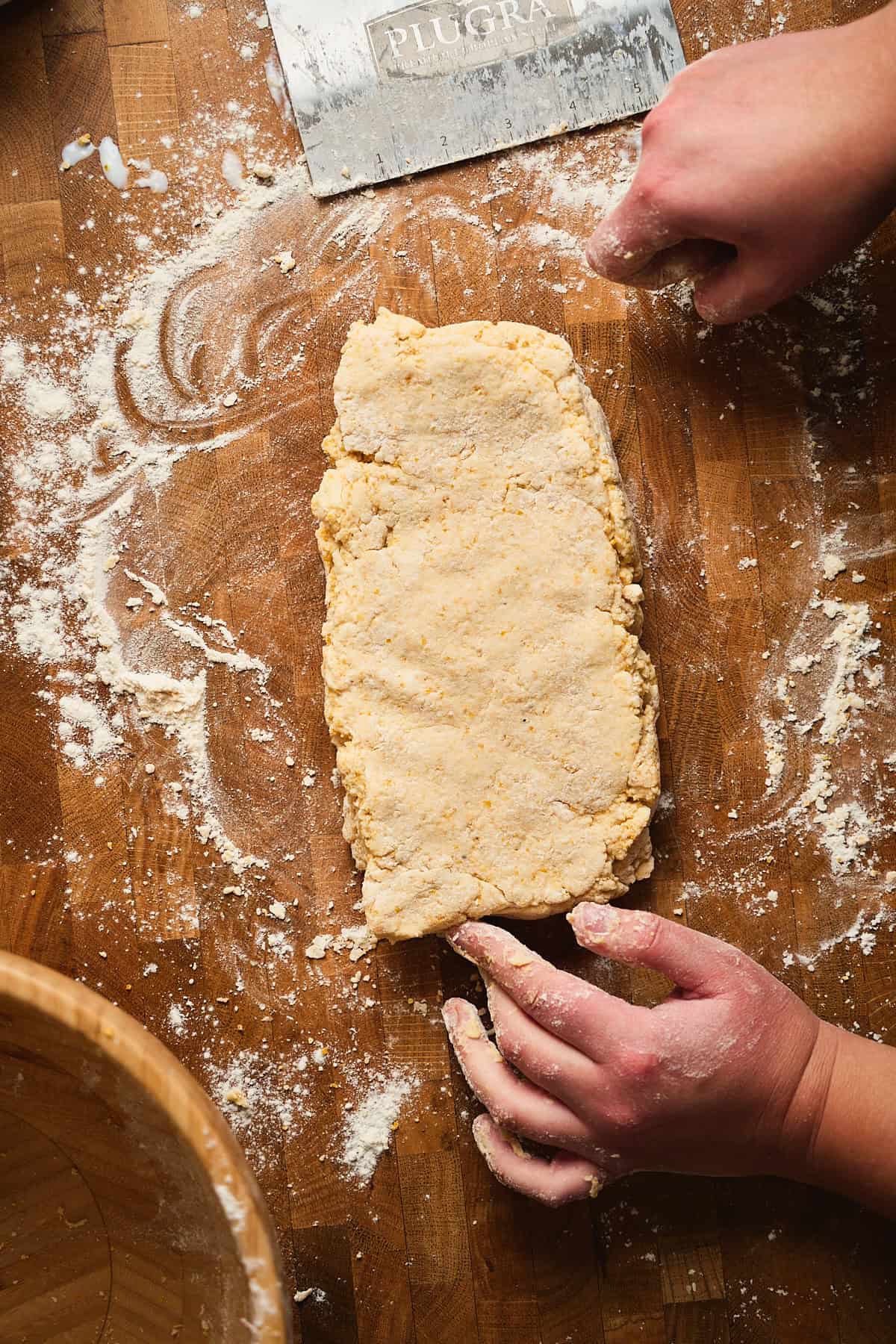 Plain dough on a wooden surface with flour, knife, and hands preparing baked goods in a cozy kitchen scene.