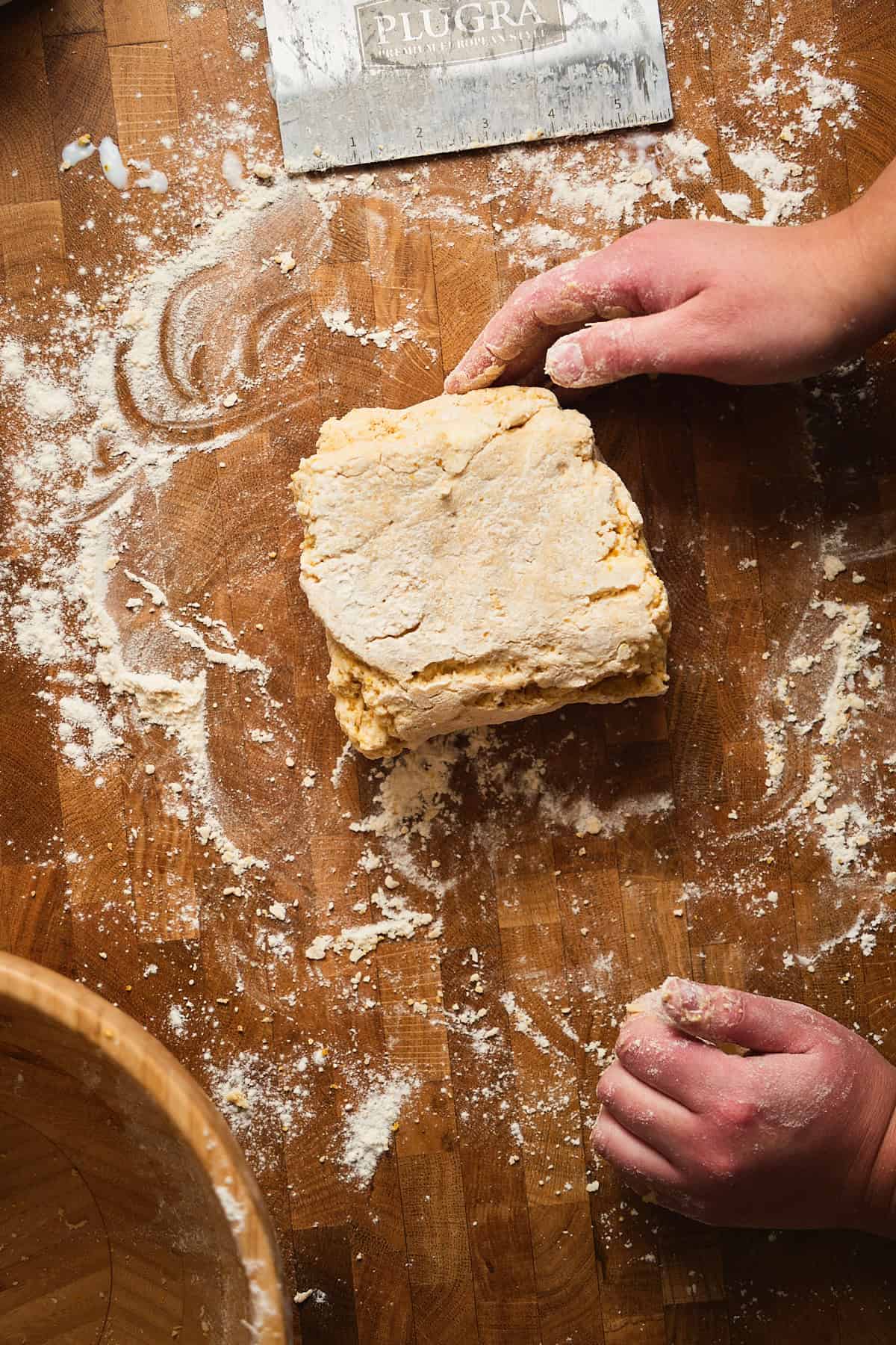 Hands pressing together leftover biscuit dough to re-roll it on a floured, wooden surface.