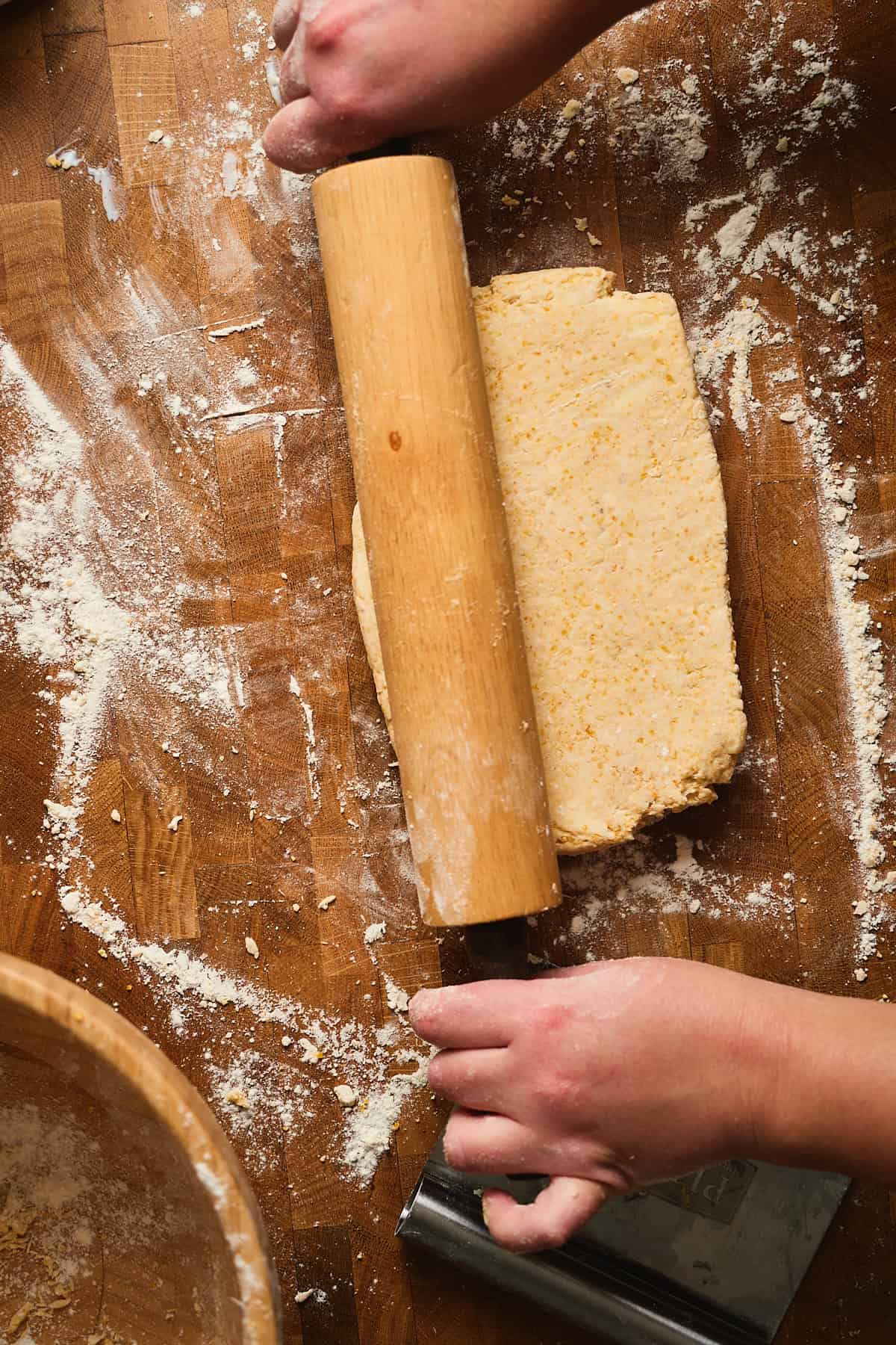 Biscuit dough being rolled out on a floured wooden surface with a rolling pin.