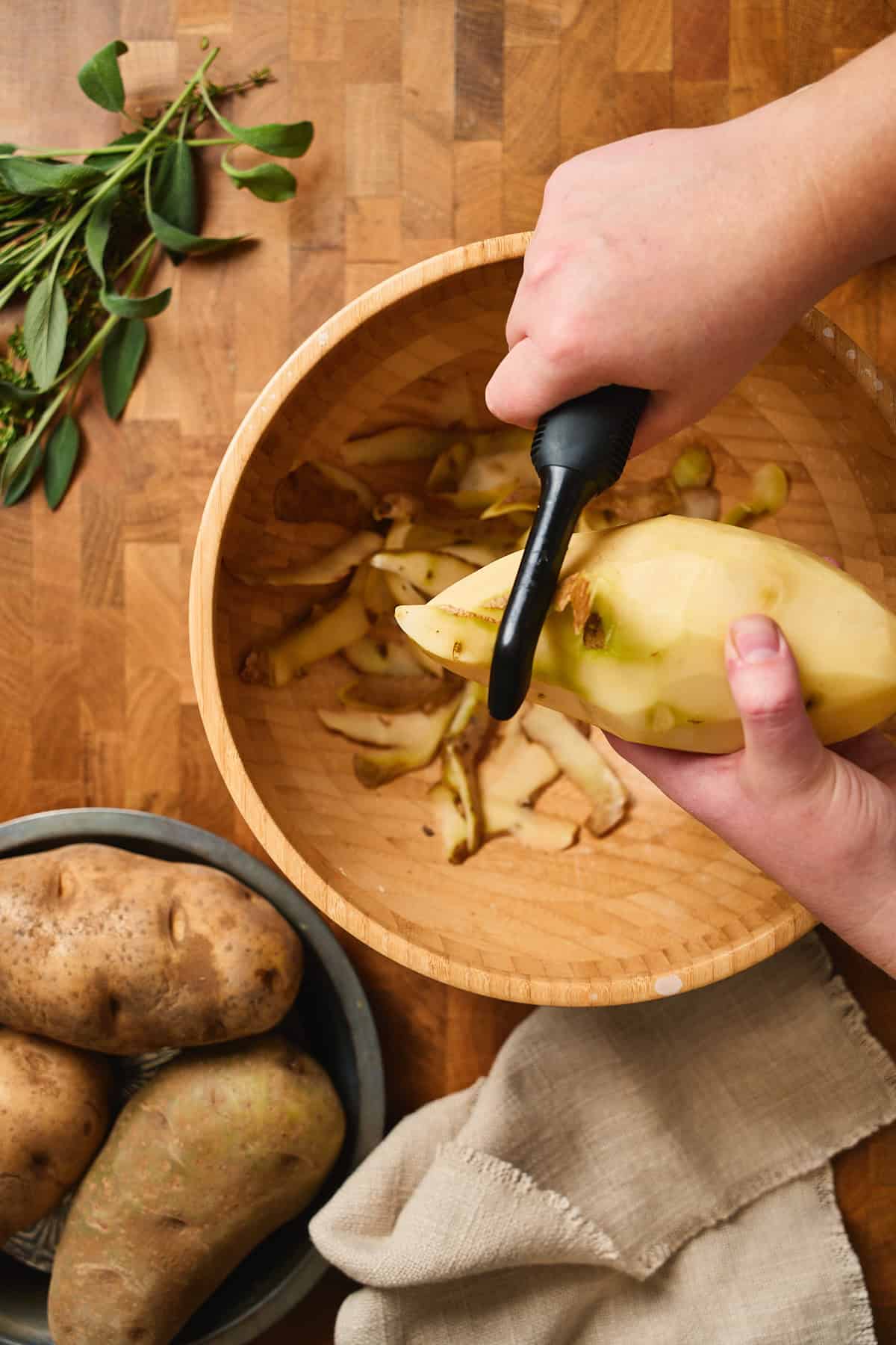Hand peeling a large russet potato with a peeler over a bowl of peels, with a stack of potatoes and fresh bundle of herbs nearby.