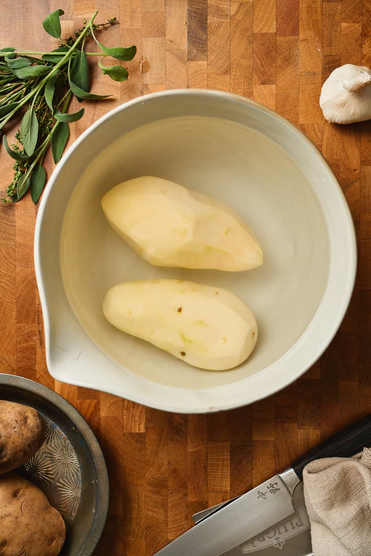 Two peeled russet potatoes in a large bowl of cold water surrounded by unpeeled potatoes, garlic, and fresh herbs.