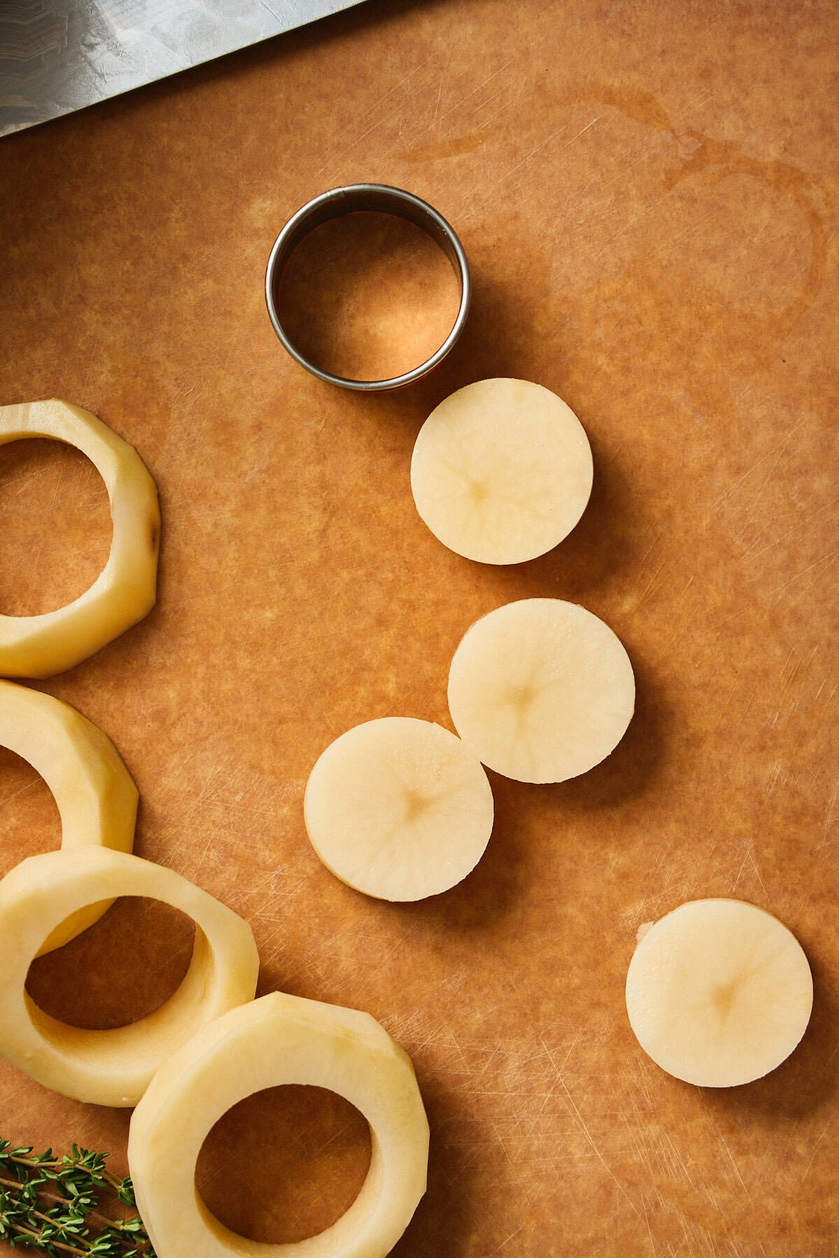 Sliced potato rounds with scraps and a cookie cutter beside herbs on a cutting board.