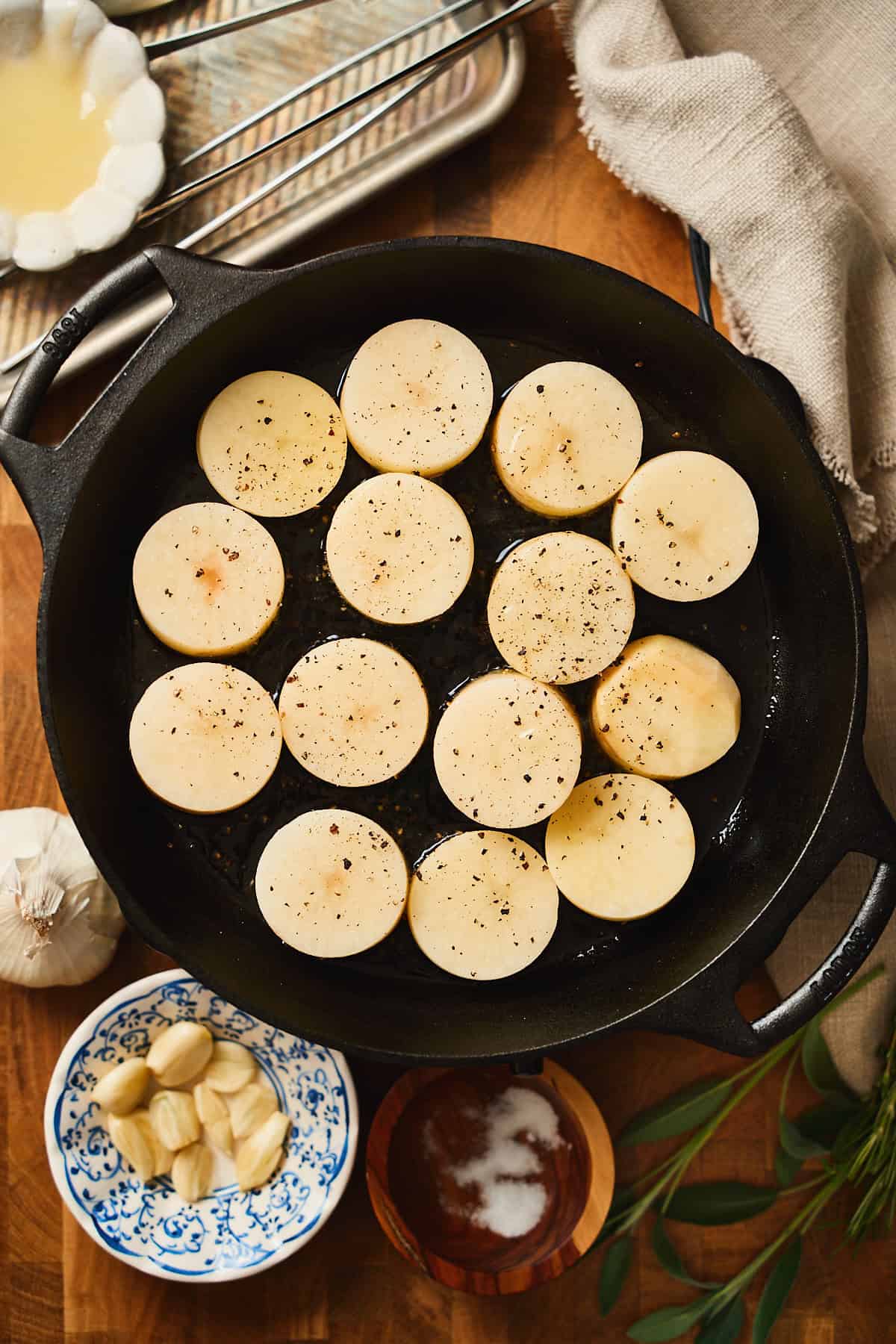 Potatoes cut into rounds in a cast iron skillet seasoned with salt and pepper, with smashed garlic cloves nearby.