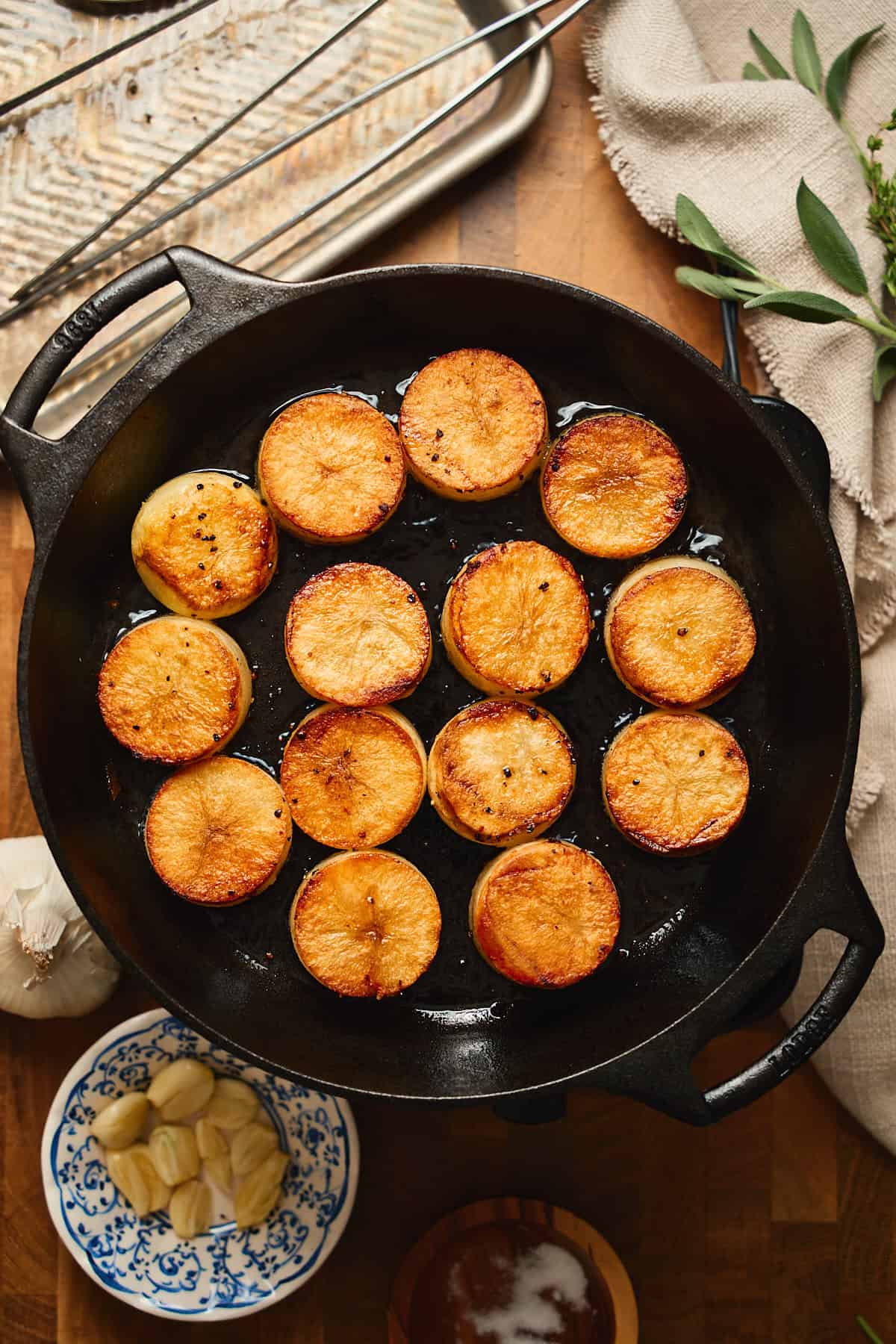 Deeply golden brown potato rounds in a cast iron skillet surrounded by garlic, fresh herbs, linens, and kitchen tools.