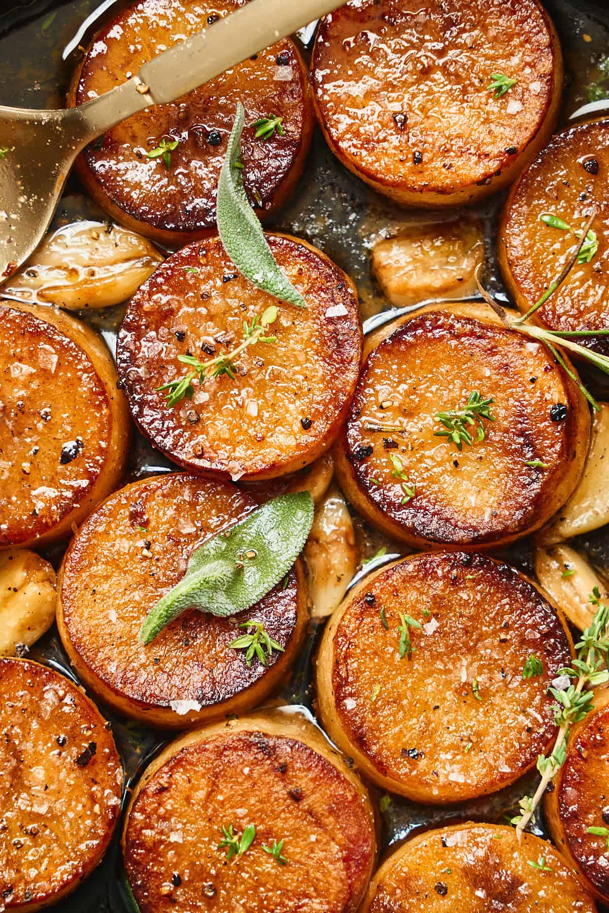 Stunning close up shot of fondant potato rounds in a pan with roasted garlic, sage leaves, fresh thyme, rosemary, and flaky salt.