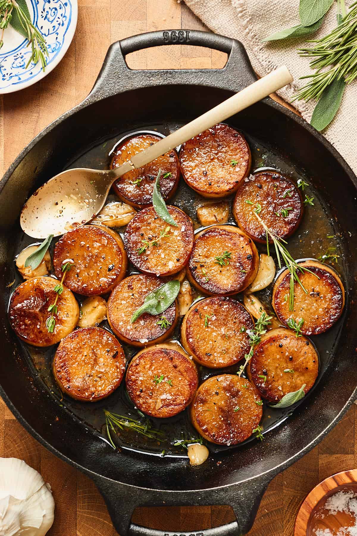 Roasted potato rounds in a buttery sauce in cast iron skillet with garlic and herbs with a golden spoon in the skillet.