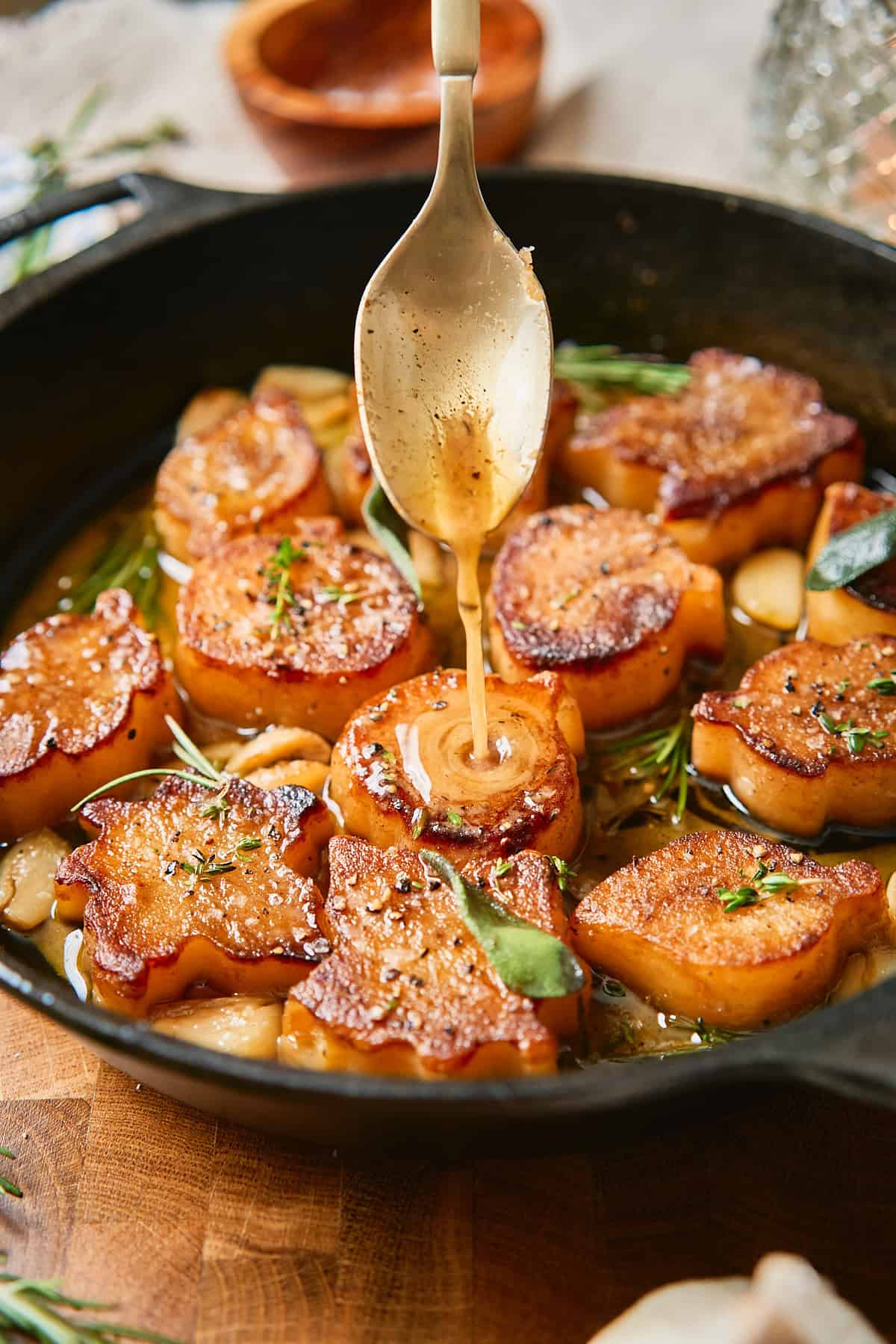Savory fondant potatoes in unique shapes being basted with a buttery white wine sauce with garlic and herbs in a cast iron skillet.