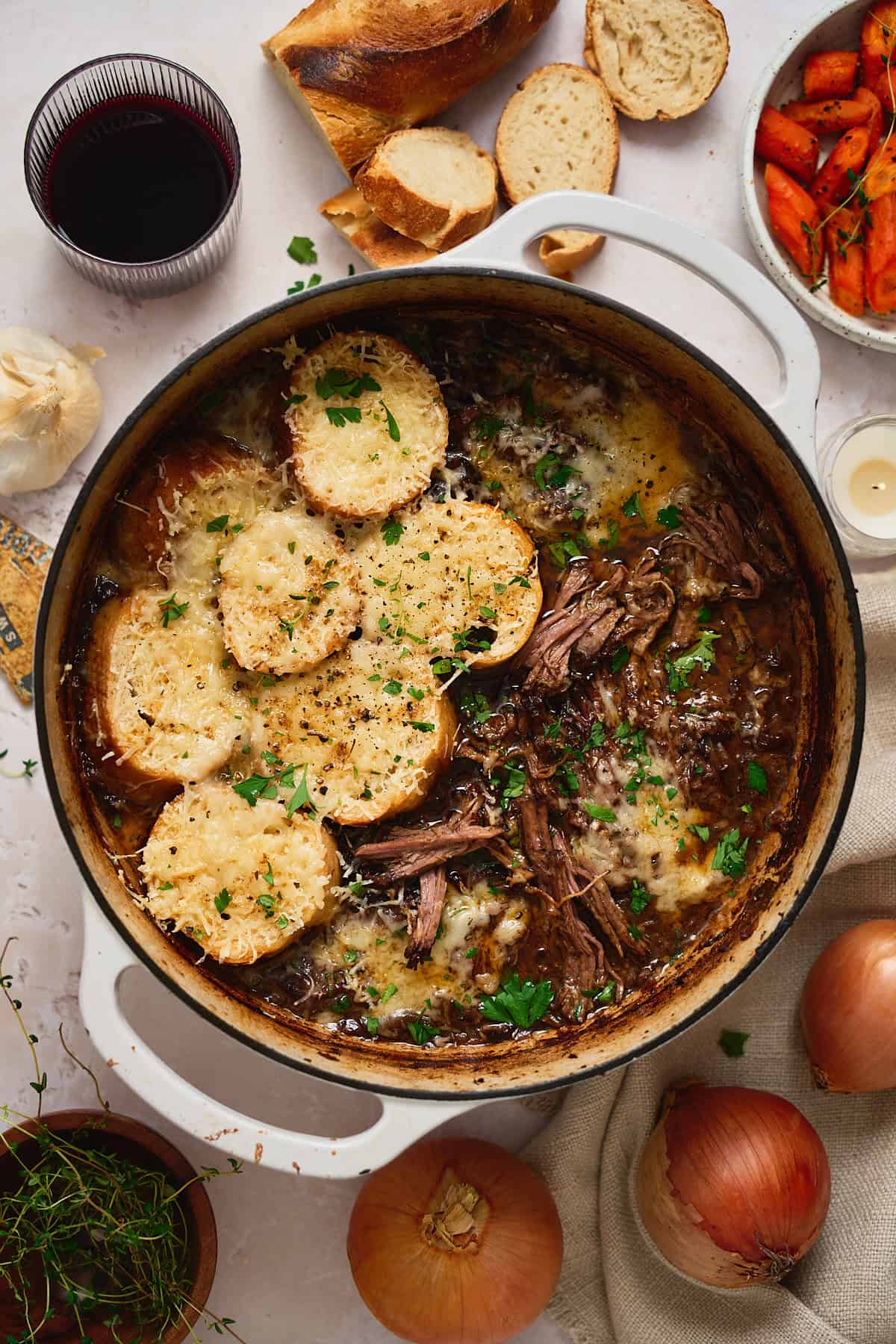 Large enameled Dutch oven filled with shredded, tender beef and topped with cheesy gruyere cheese toasts, and herbs surrounded by wine, garlic, onions, and more ingredients used to make the roast.