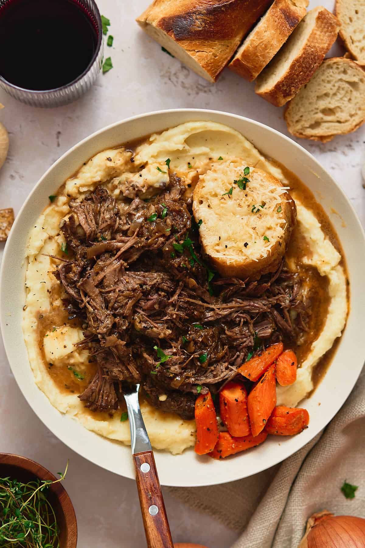 Bowl of creamy mashed potatoes topped with tender, shredded beef, roasted carrots, pan gravy, and cheesy gruyere toast.