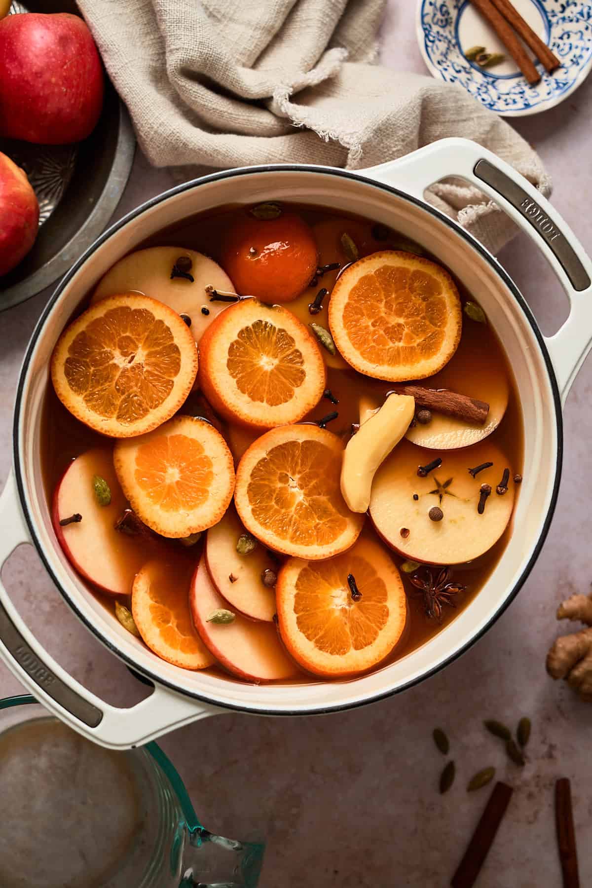 Fresh orange slices, apple pieces, and spices in a Dutch oven mulling together surrounded by other ingredients.
