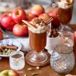 Creamy apple-cinnamon spiced beverage topped with whipped cream and cinnamon sticks, surrounded by lots of apples, whole spices, and candles.