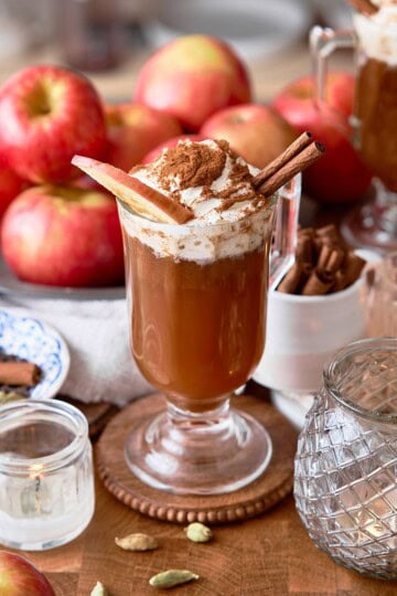 Creamy apple-cinnamon spiced beverage topped with whipped cream and cinnamon sticks, surrounded by lots of apples, whole spices, and candles.