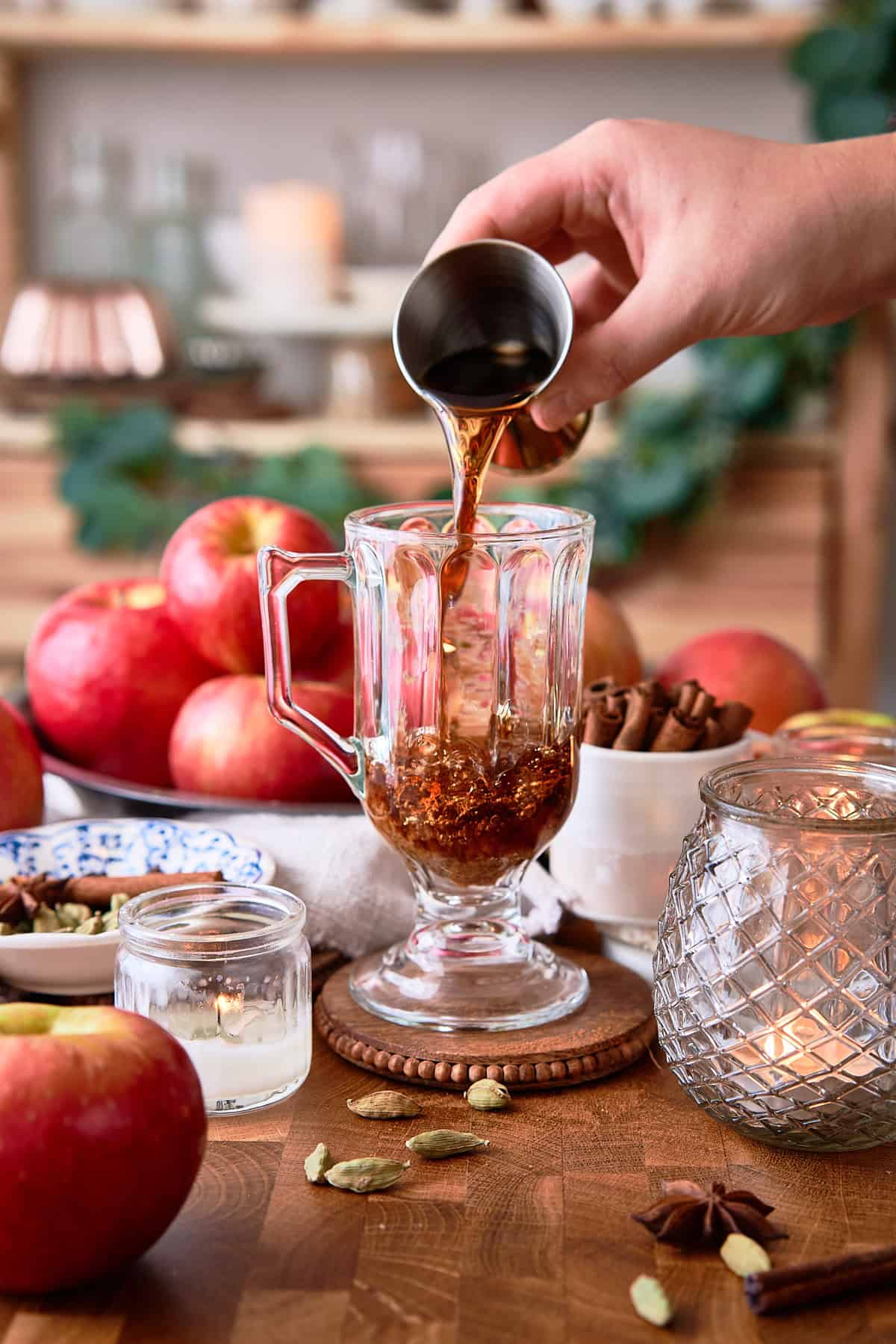 Dark rum being poured into a glass mug with apples and spices on a wooden table.