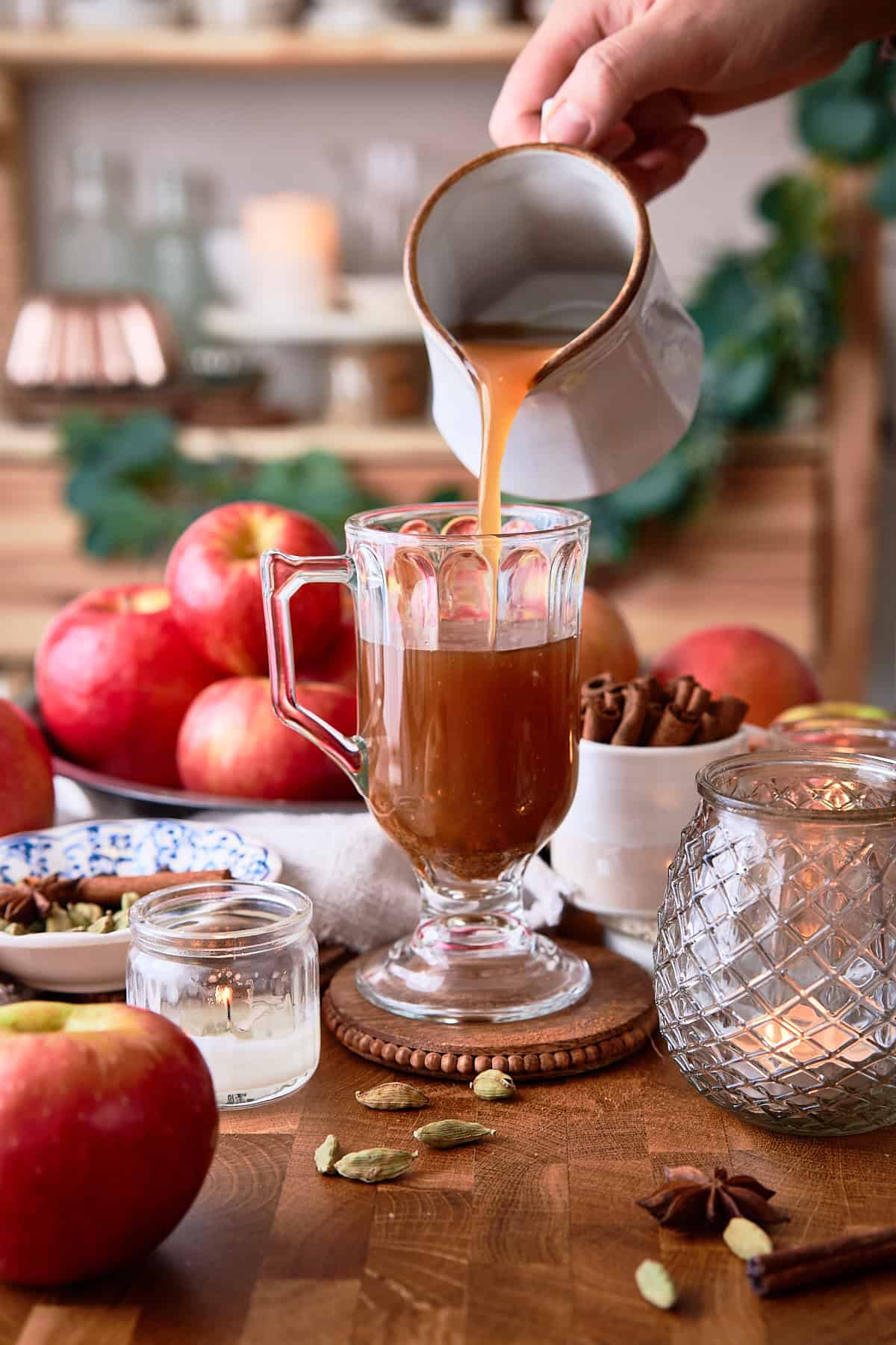 Warm apple cider being poured into a glass mug on a rustic wooden table with apples and spices.
