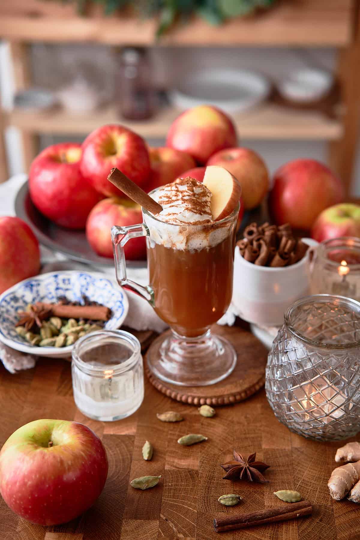 A cozy scene featuring homemade apple cider topped with whipped cream, cinnamon, and apple slices, with fresh apples and spices on a wooden table.