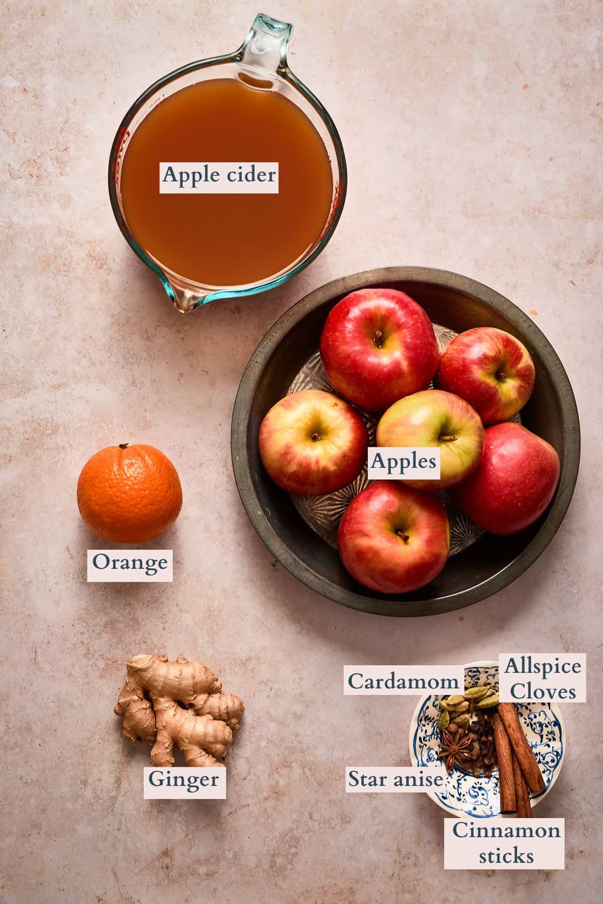Ingredients to make mulled apple cider on a tan marble backdrop labeled to denote each ingredient.