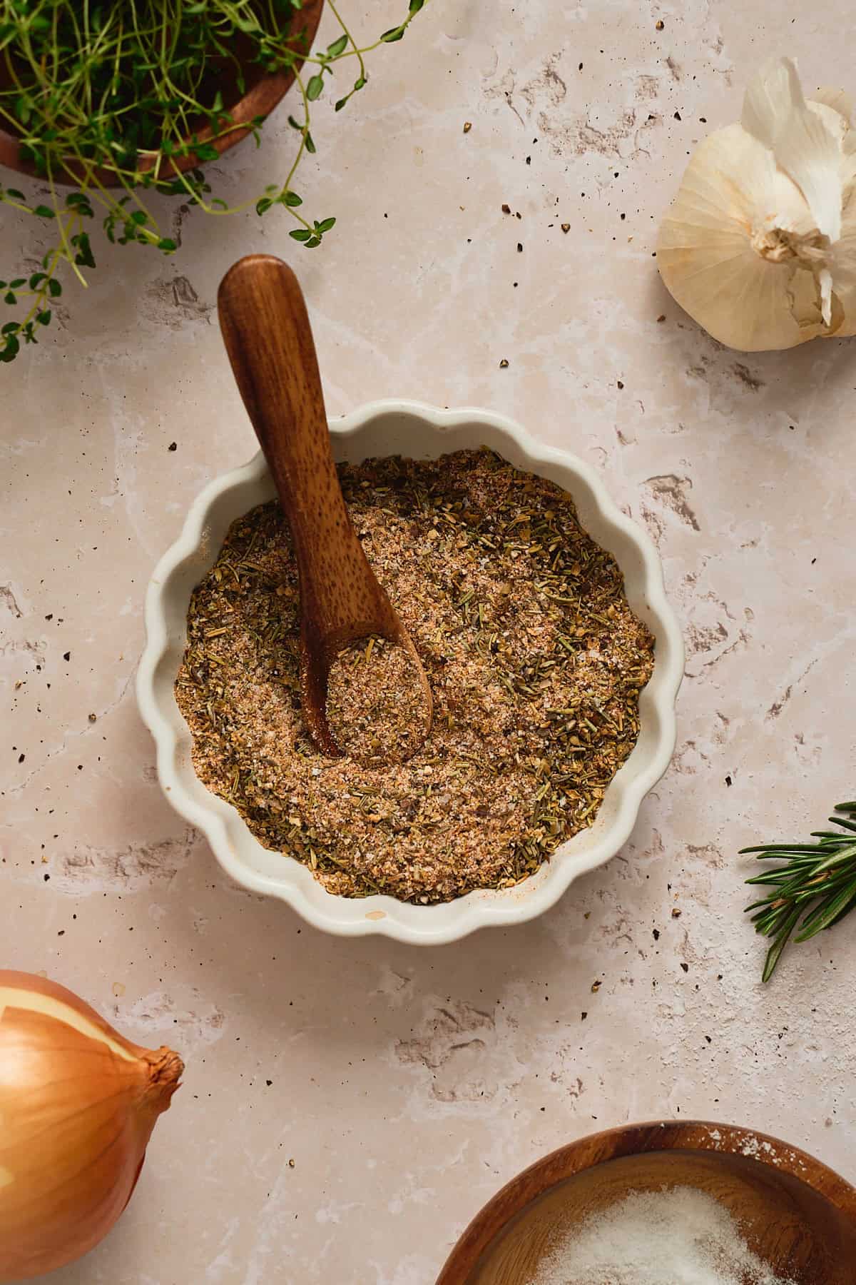 Small ruffled white bowl of homemade pot roast seasoning with a small wooden spoon inside, surrounded by onion, garlic, thyme, rosemary, and salt.