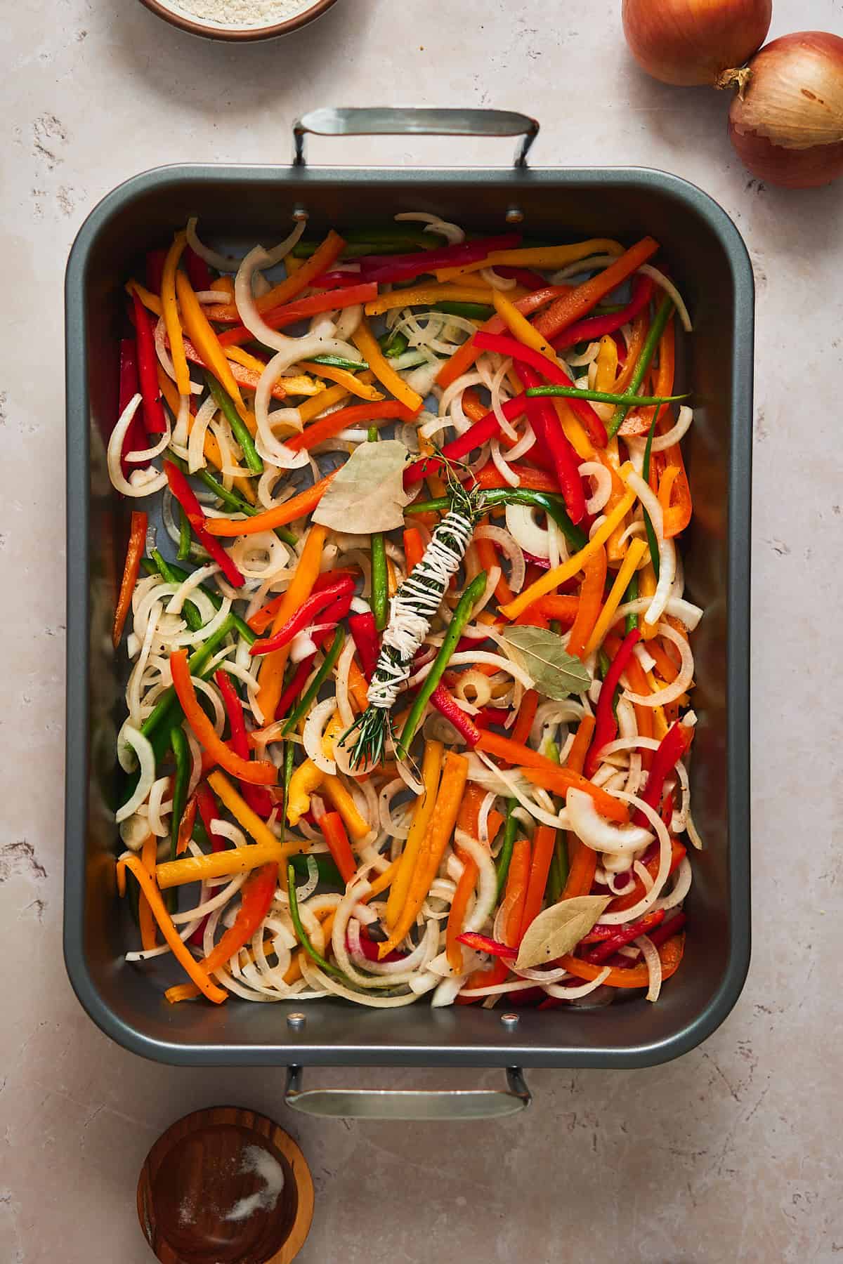 Bell peppers and onions with bay leaves and an herb bundle on the bottom of a large roasting pan.
