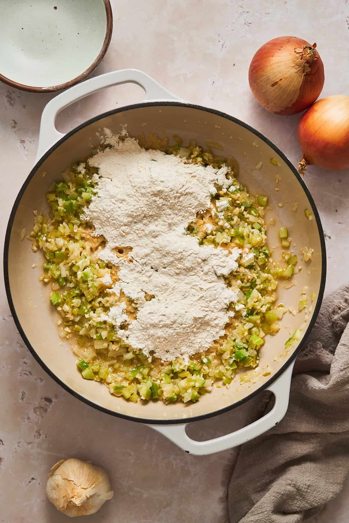 Onions, celery, and garlic cooking in a pan of hot butter with flour added to the top.