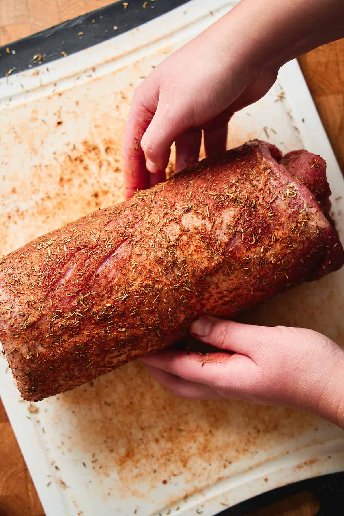 Tightly rolled pork loin roast seasoned well, with a person's two hands tucking the roast into a round log shape.