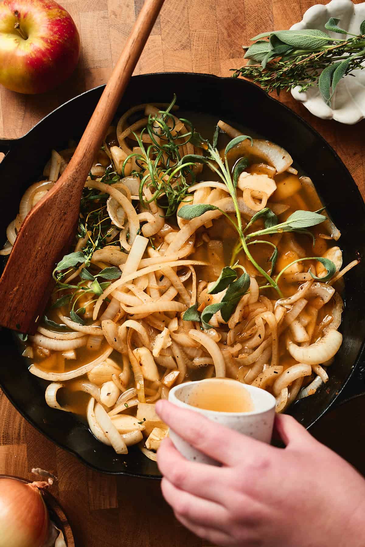 Hand pouring liquid into a skillet with cooked onions, garlic, and herbs.