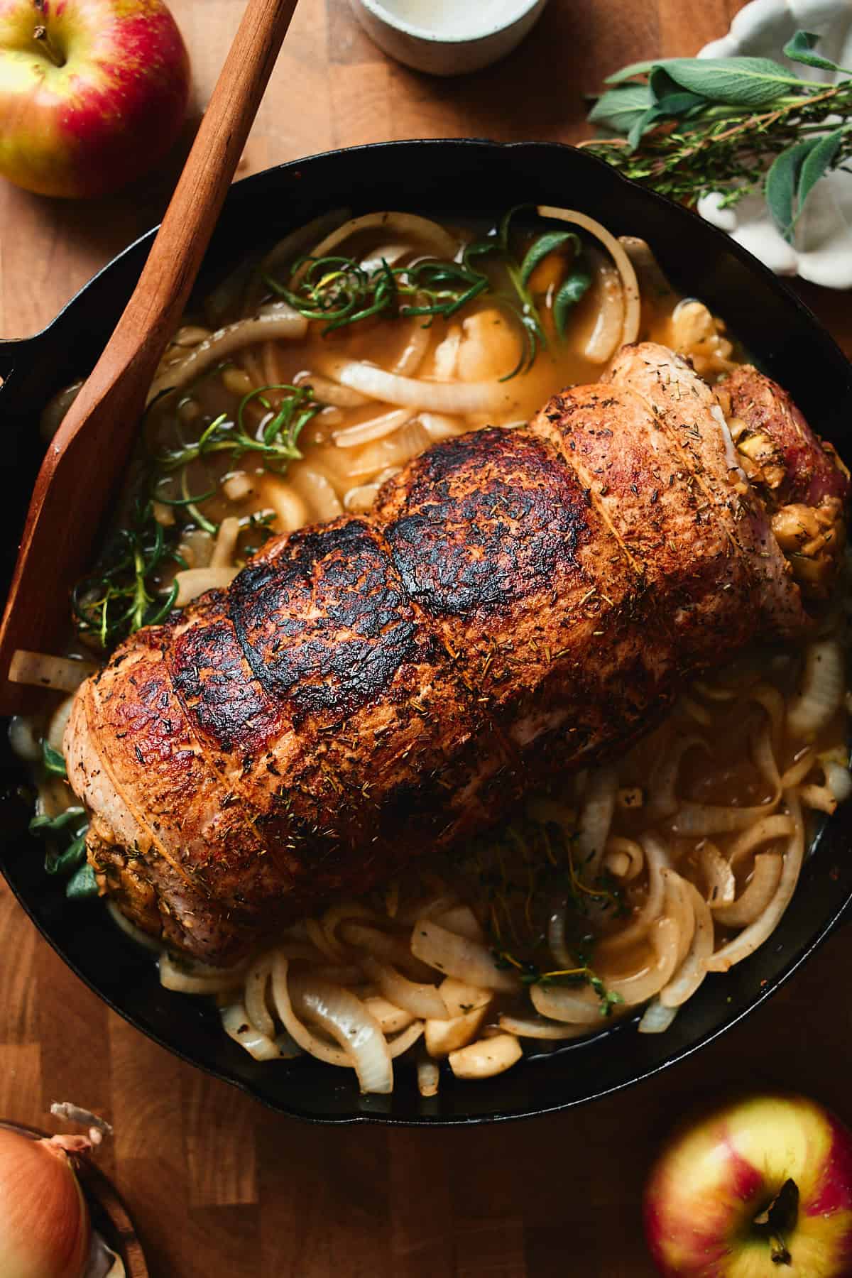 Seared pork loin roast on top of a bed of onions, herbs, and garlic in a deep cast iron skillet.