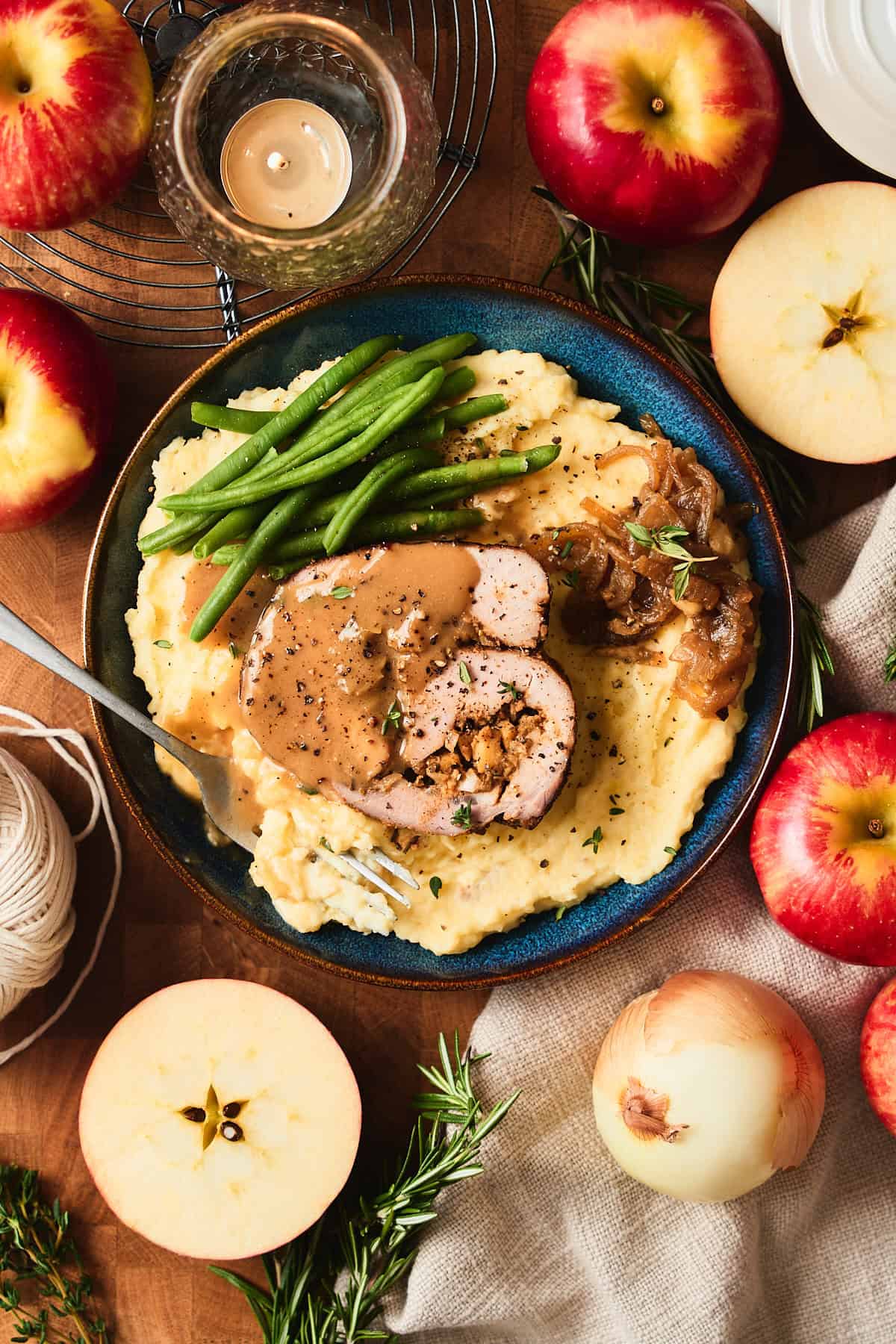 Beautiful pork loin stuffed with apple filling and rolled into a pinwheel shape on a bed of mashed potatoes with green beans and gravy surrounded by apples, herbs, and candles.