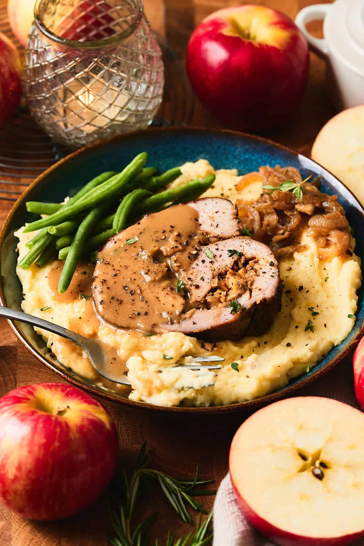 Gorgeous stuffed pork loin spiral on a bed of mashed potatoes with onions, green beans, and apple cider gravy, surrounded by fresh apples, herbs and candles.