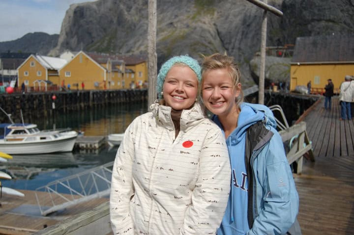 Two women standing in a small Norwegian fishing village with fjords in the background.