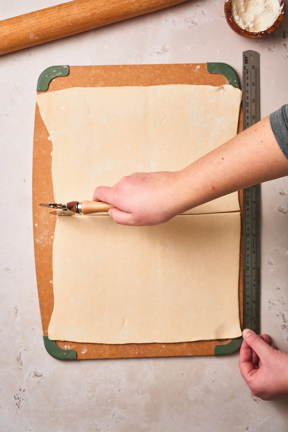 Hands cutting a large sheet of puff pastry in half with a ruler and pastry wheel.