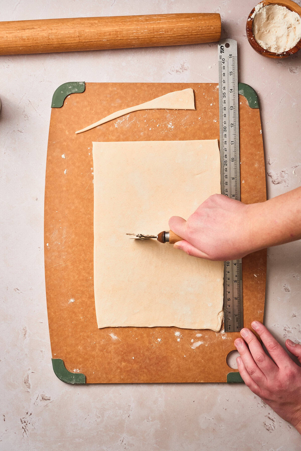 Hand cutting a sheet of puff pastry in half with a ruler, and pastry wheel with scraps of dough nearby.