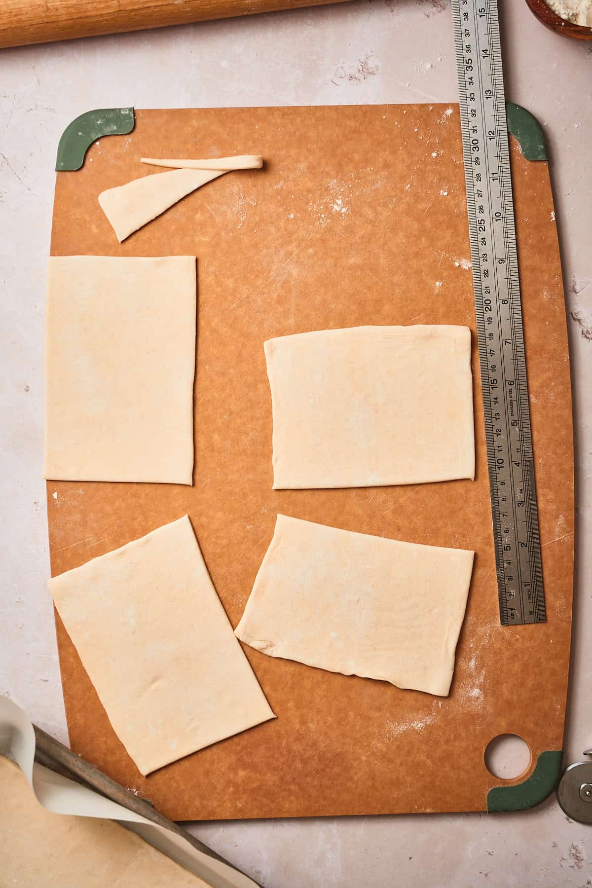 Four roughly cut squares of puff pastry with a ruler and pastry scarps nearby.