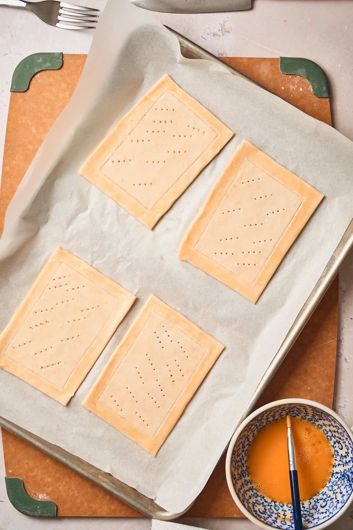 Prepped puff pastry squares on a parchment lined baking sheet, with the edges brushed with nearby eggwash.