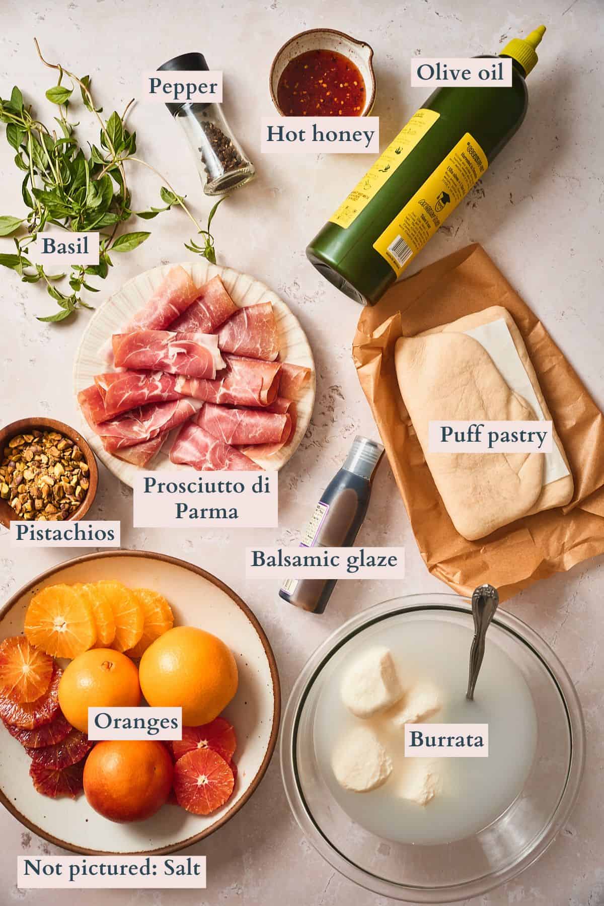Ingredients to make burrata and prosciutto puff pastry tarts laid out on a table with oranges, basil and other ingredients and labeled.