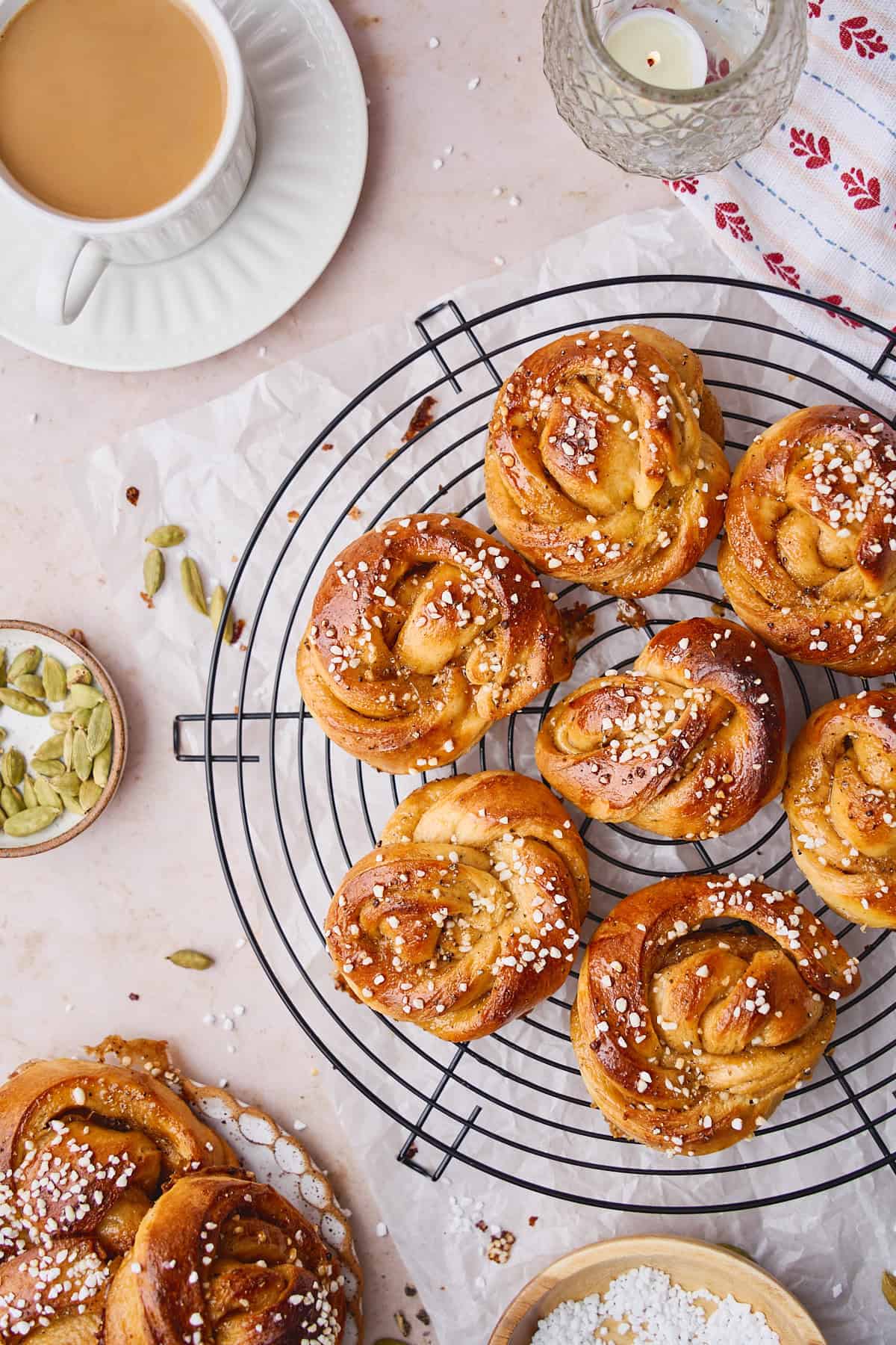 Flat lay of baked cardamom buns topped with pearl sugar on a round cooling rack with coffee and green cardamom pods nearby.
