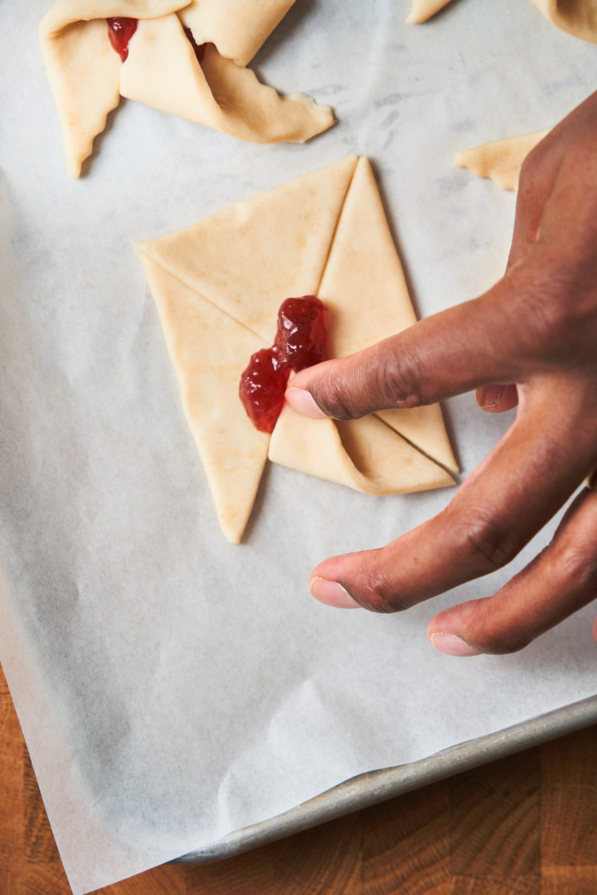 Hand folding pieces of puff pastry corners over the center of a jam filling to create a pinwheel shape.