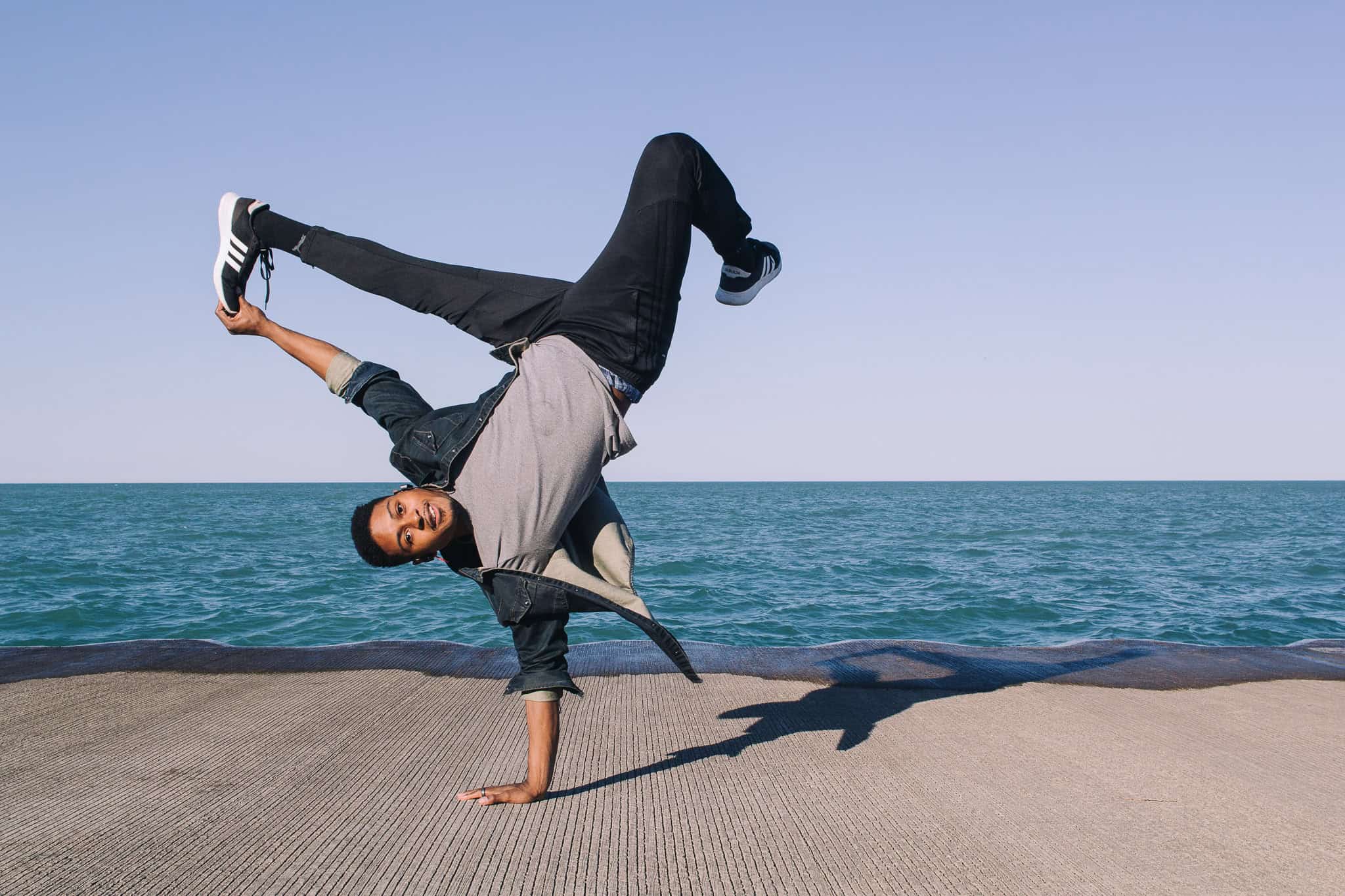 Man breakdancing in front of water on pavement doing a freeze holding his leg.
