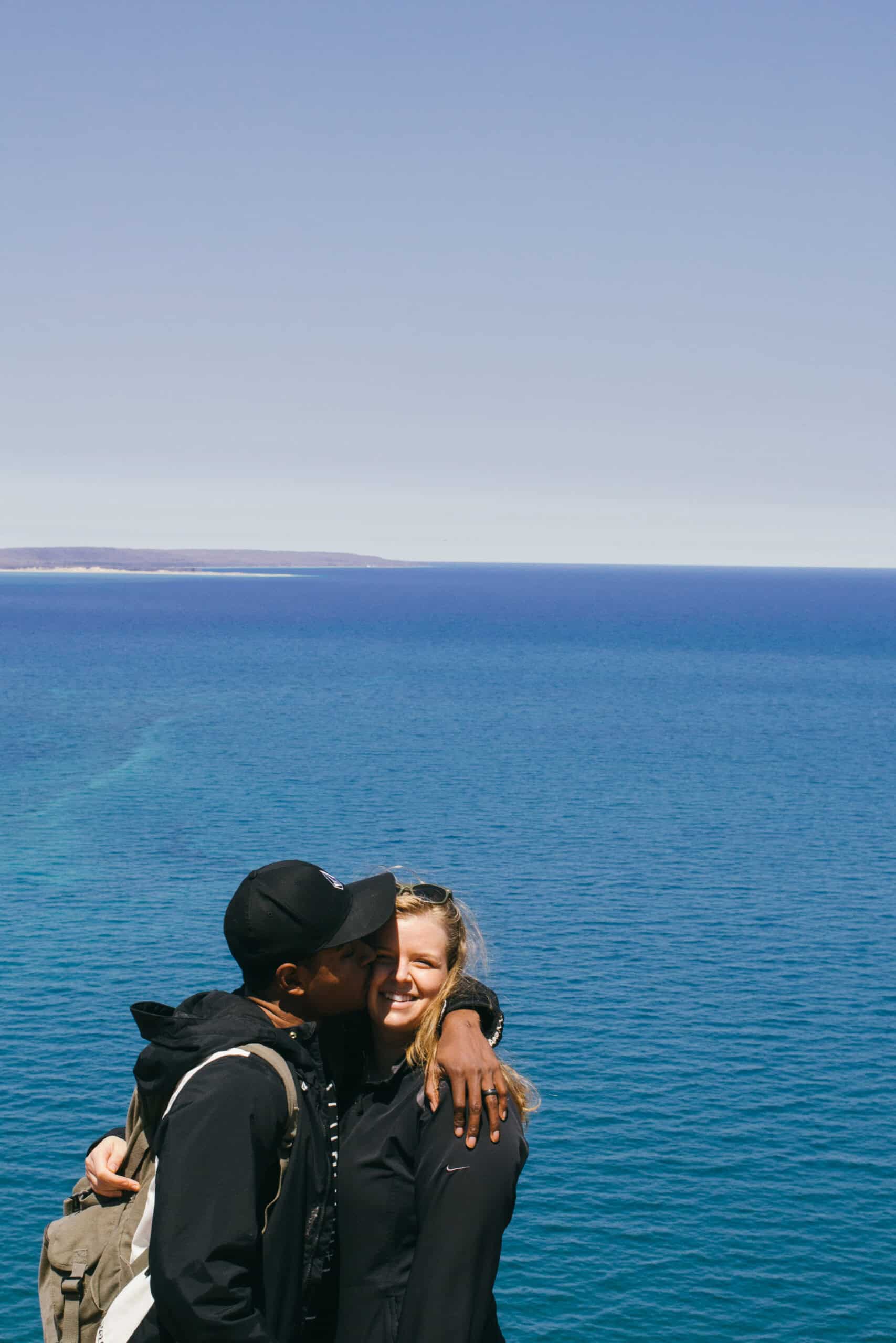 man and woman in front of a beautiful huge body of blue water on a tall sand dune, and the man is giving the woman a kiss on the cheek.