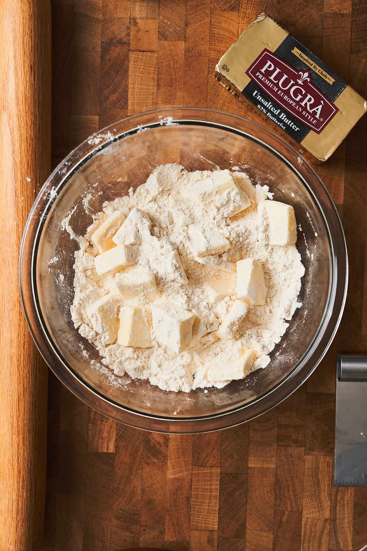 Chunks of butter coated in flour in a bowl on a wooden surface with a block of Plugra nearby.