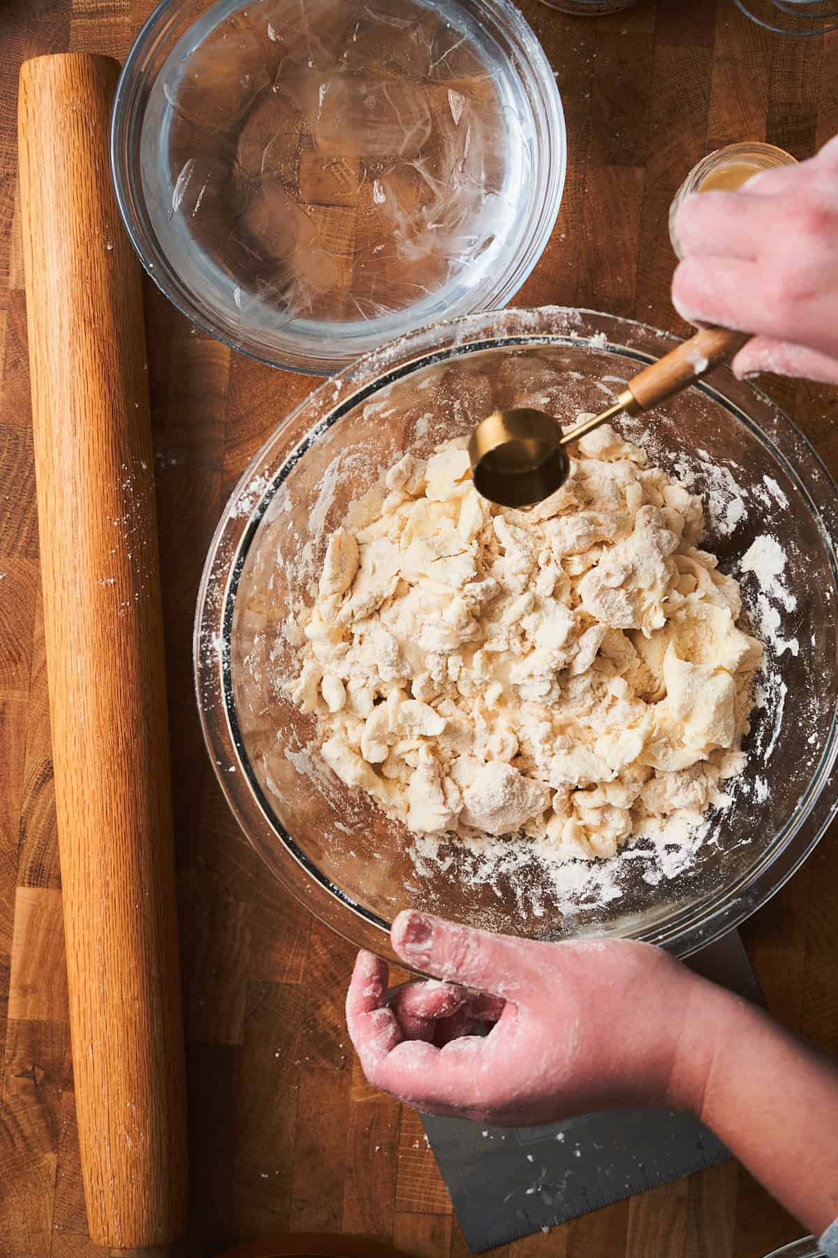 Hand adding 1 tablespoon of water at a time to a bowl of flour and butter sheets to make puff pastry dough.