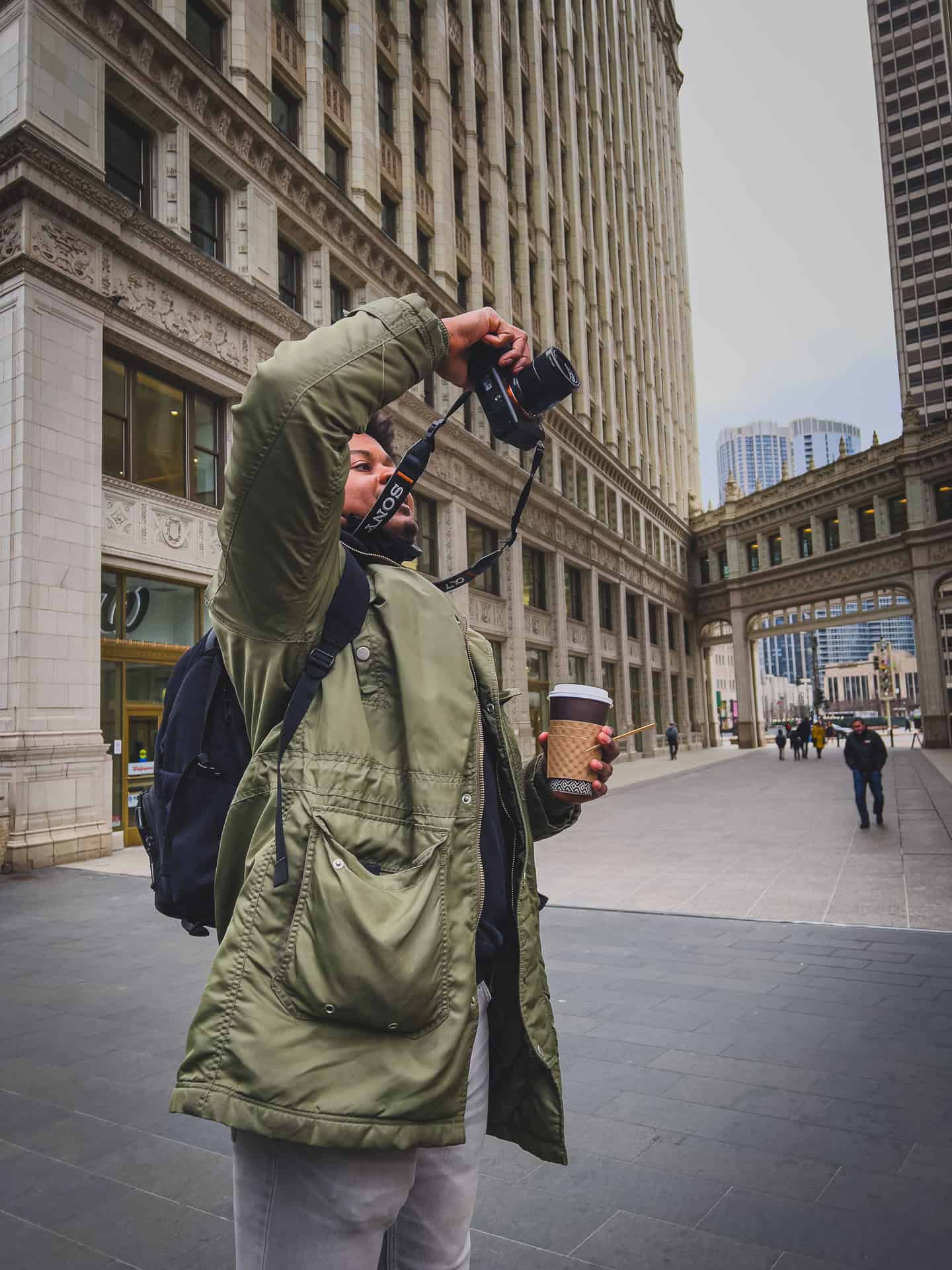 A man in a green jacket holding a coffee reaching over his head to take a photo in downtown Chicago.
