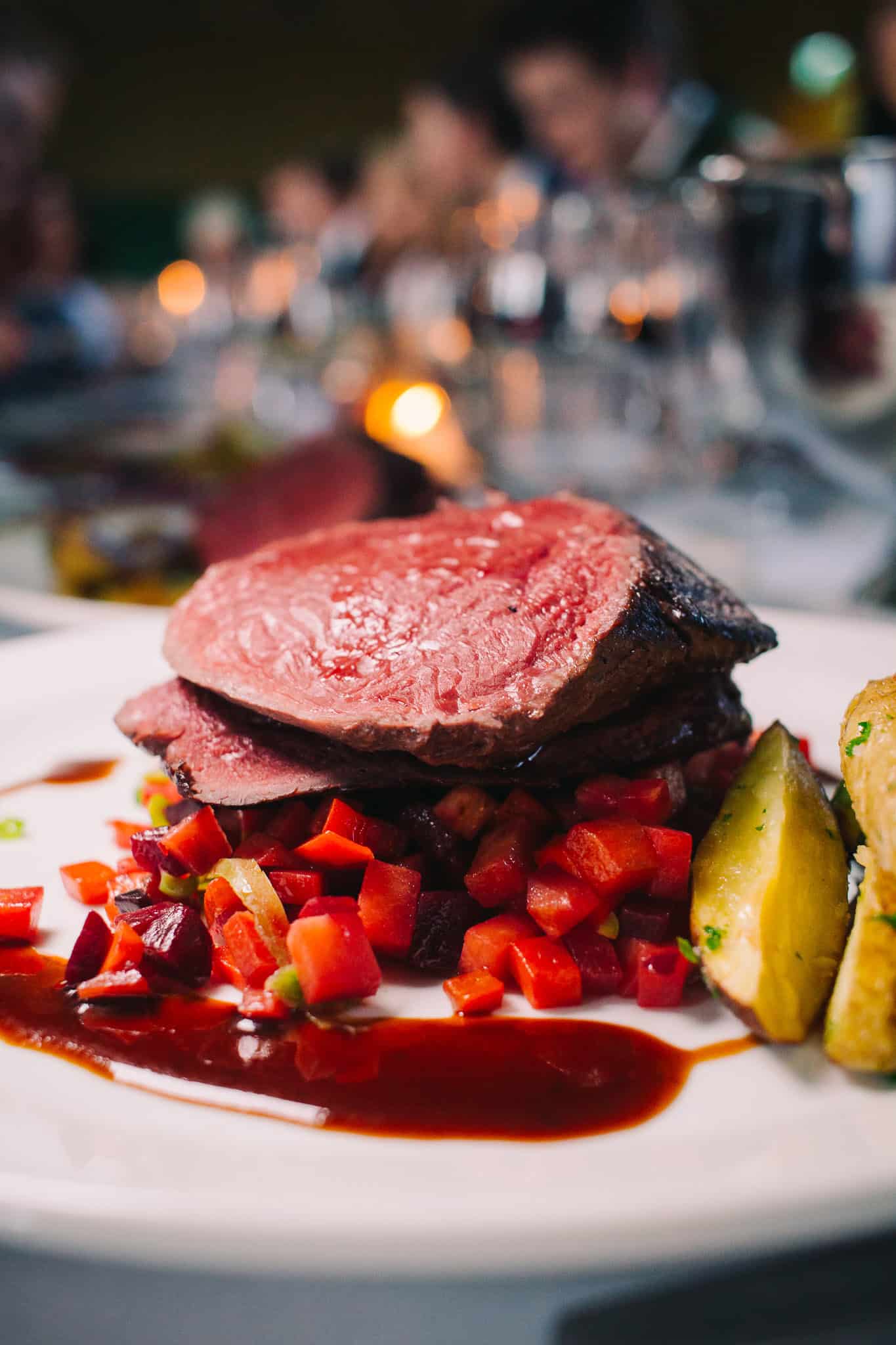 Photo of a gorgeous medium rare reindeer steak with veggies, potatoes, and lingonberry sauce on a table with many guests in the background.