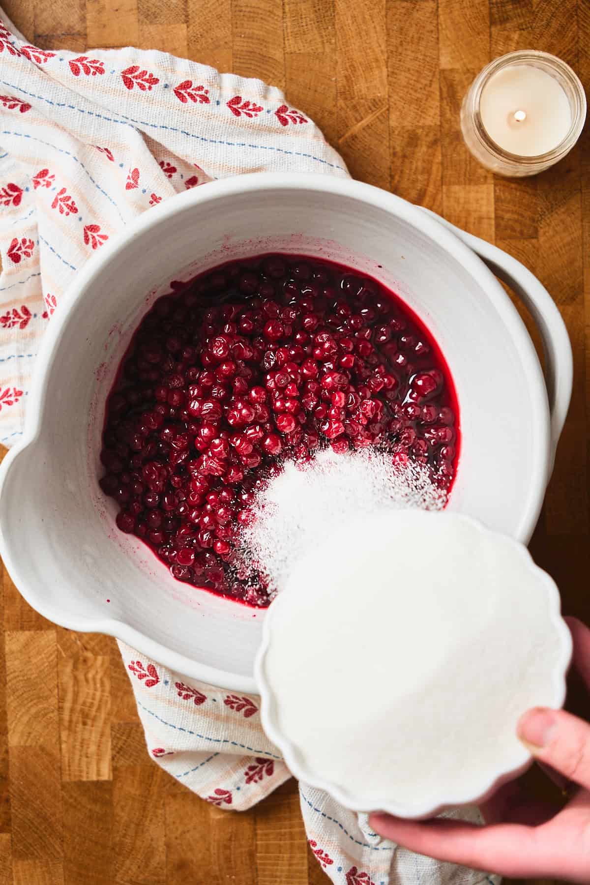 Hand pouring sugar into lingonberries in a bowl.