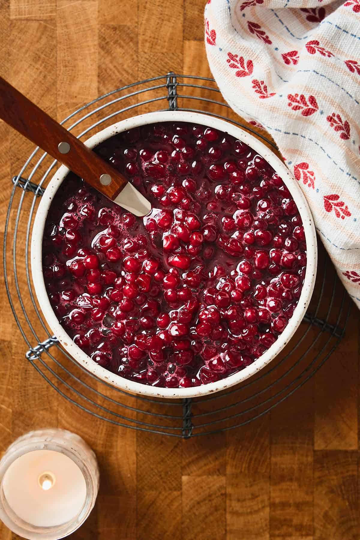 Bowl of bright red gorgeous lingonberries on a wooden surface, with a Scandi linen nearby, and a candle on the scene.