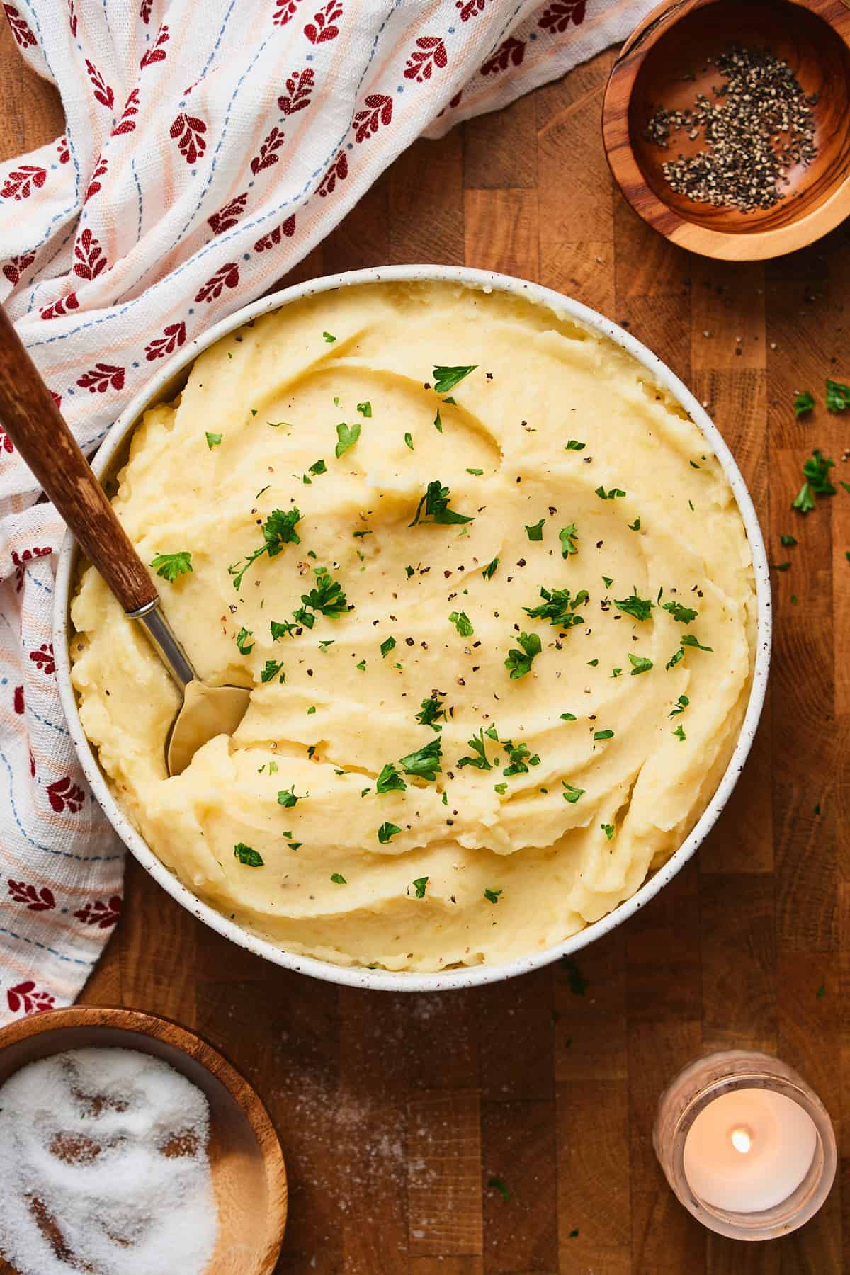 Bowl of mashed potatoes topped with parsley on a wooden backdrop with candles surrounding and cute Nordic printed linen.
