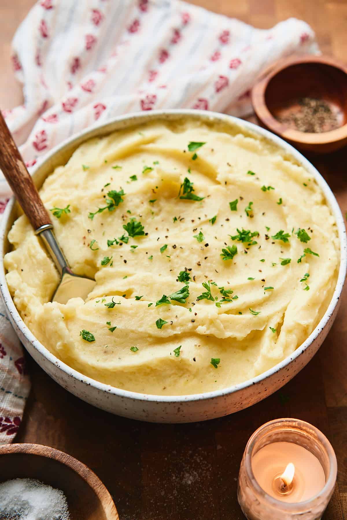 45 degree angle of a big bowl of mashed potatoes topped with parsley, with a cute Nordic printed napkin nearby and a candle up front.